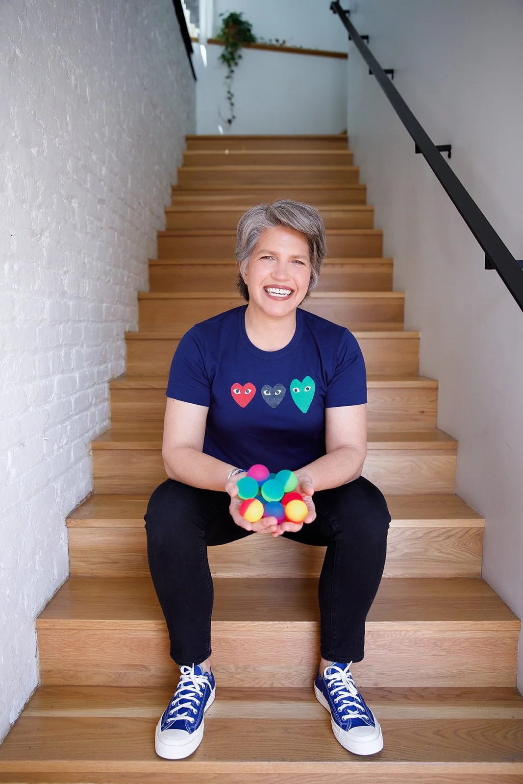 A smiling woman with short gray hair sitting on wooden stairs, holding colorful glow-in-the-dark balls.