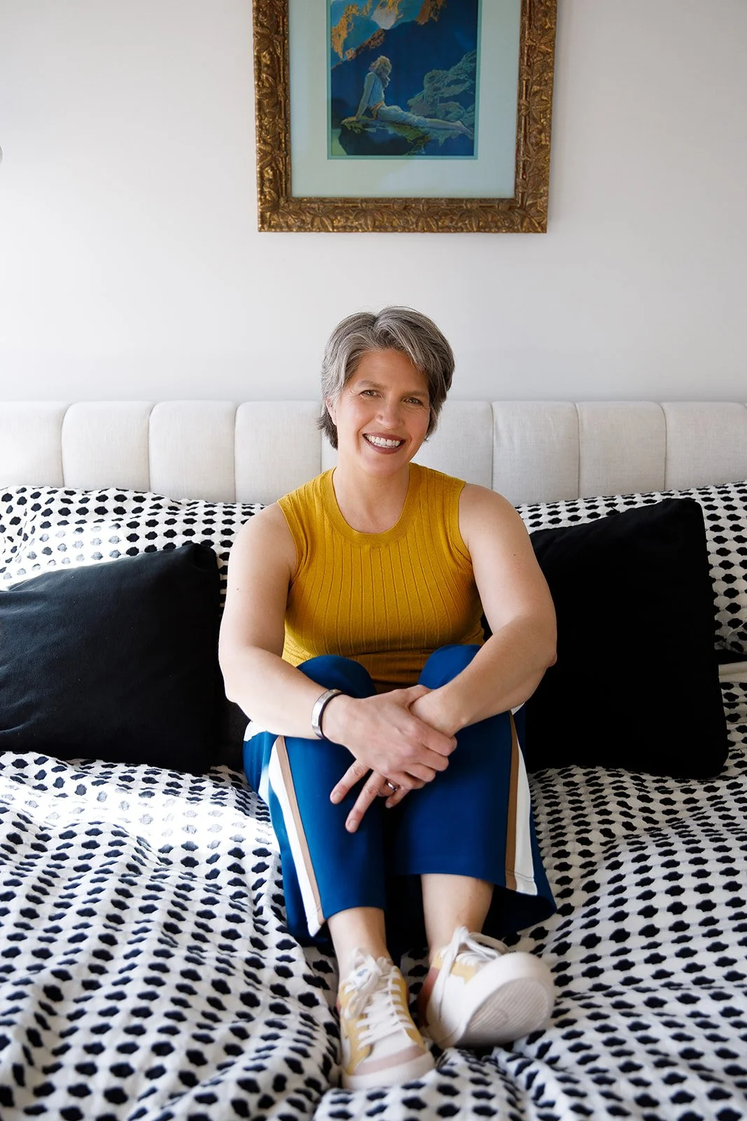 A woman with short gray hair sitting on a bed, smiling at the camera, wearing a yellow sleeveless top, blue pants, and sneakers, with a framed painting hanging on the wall behind her.