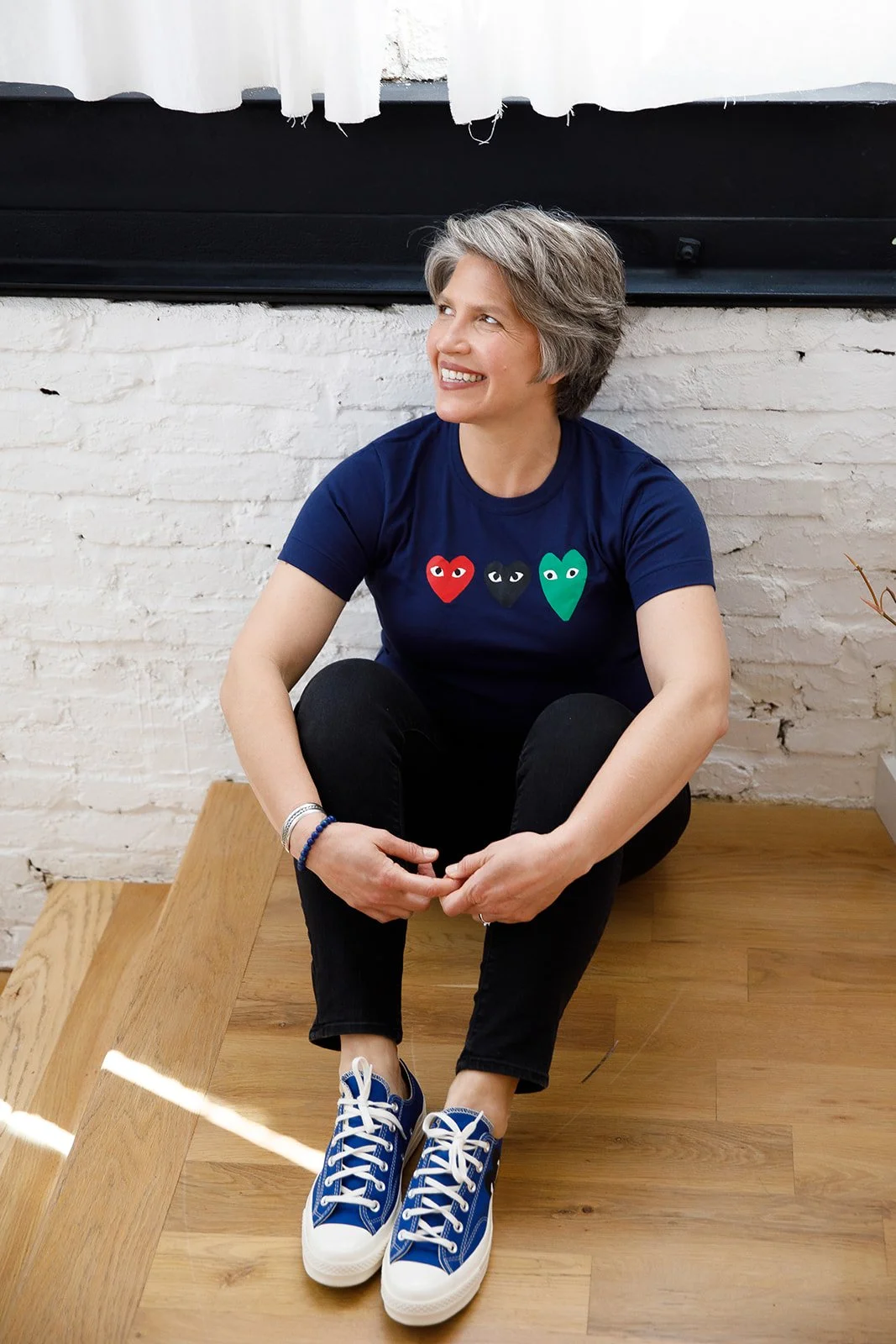 Older woman with short gray hair sitting on a wooden floor against a white painted brick wall. She is smiling, wearing a navy blue T-shirt with three colored heart shapes with eyes, black pants, and blue sneakers, with her hands resting on her knees.