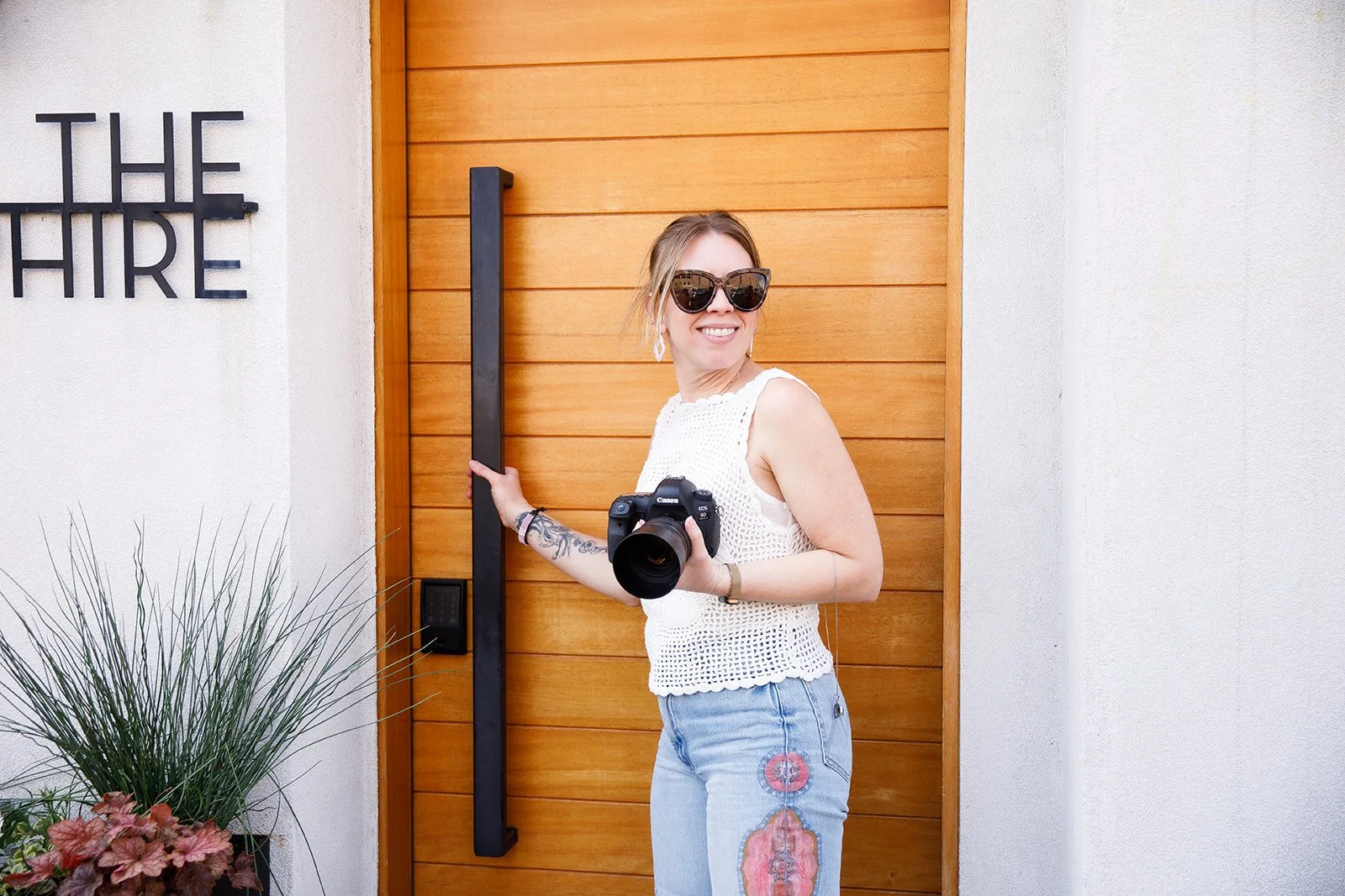 A woman standing in front of a wooden door with a camera in her hand, wearing sunglasses, a white sleeveless top, and jeans, smiling.