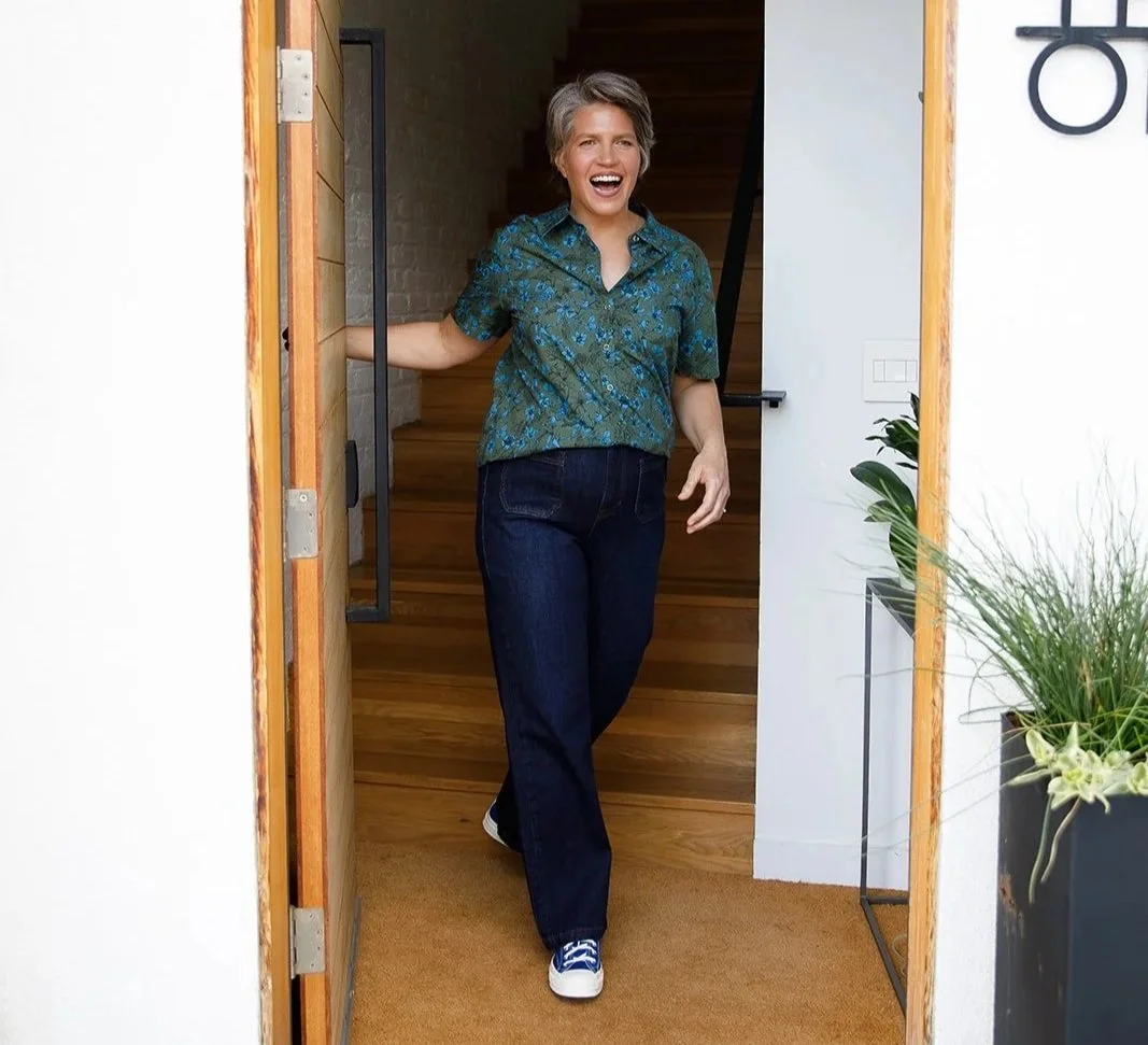 A woman with short gray hair smiling and walking through a doorway into a house, wearing a green patterned shirt, dark jeans, and sneakers.