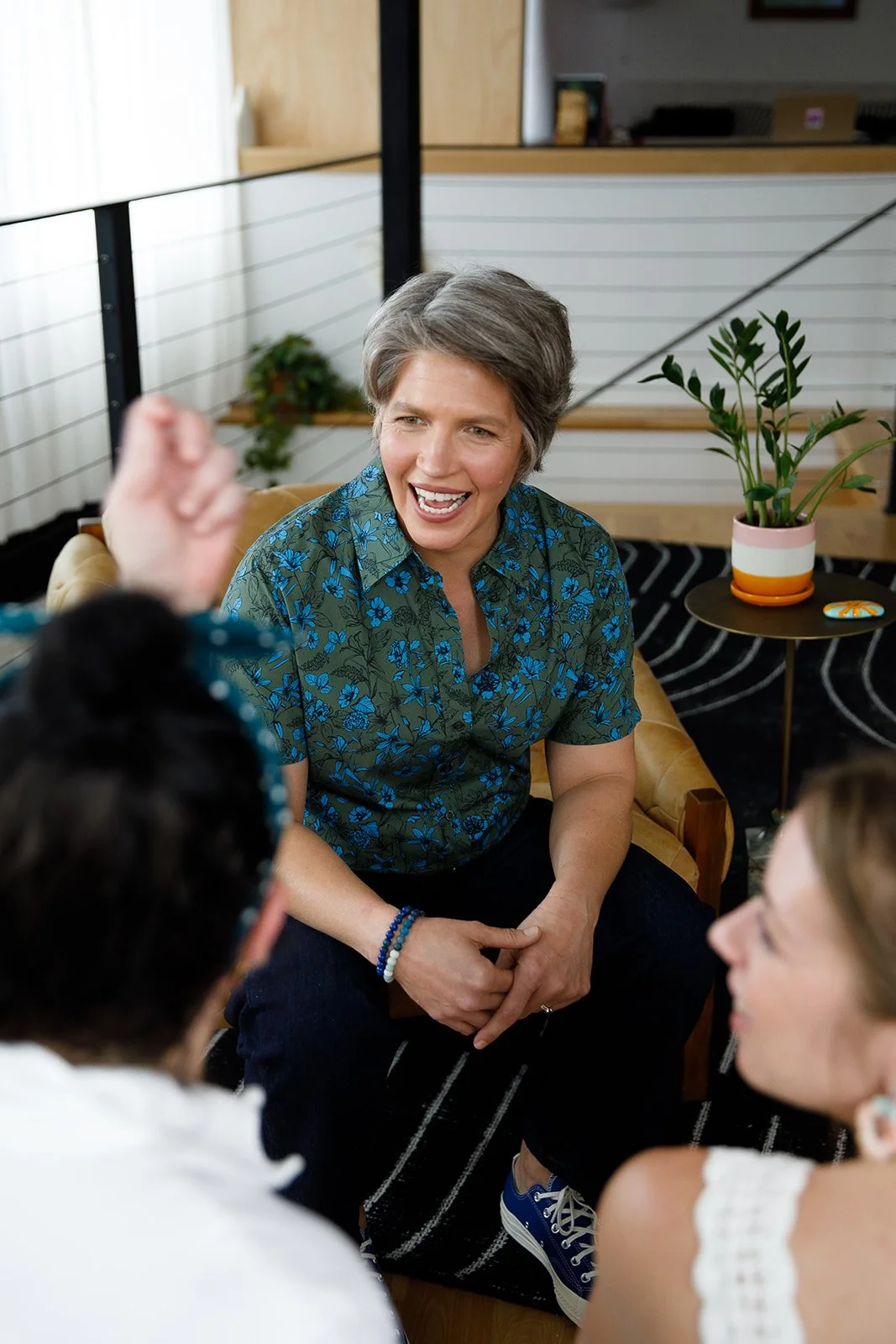 A woman with short gray hair wearing a blue floral shirt, sitting indoors on a tan chair, smiling and engaging in conversation with two other people.