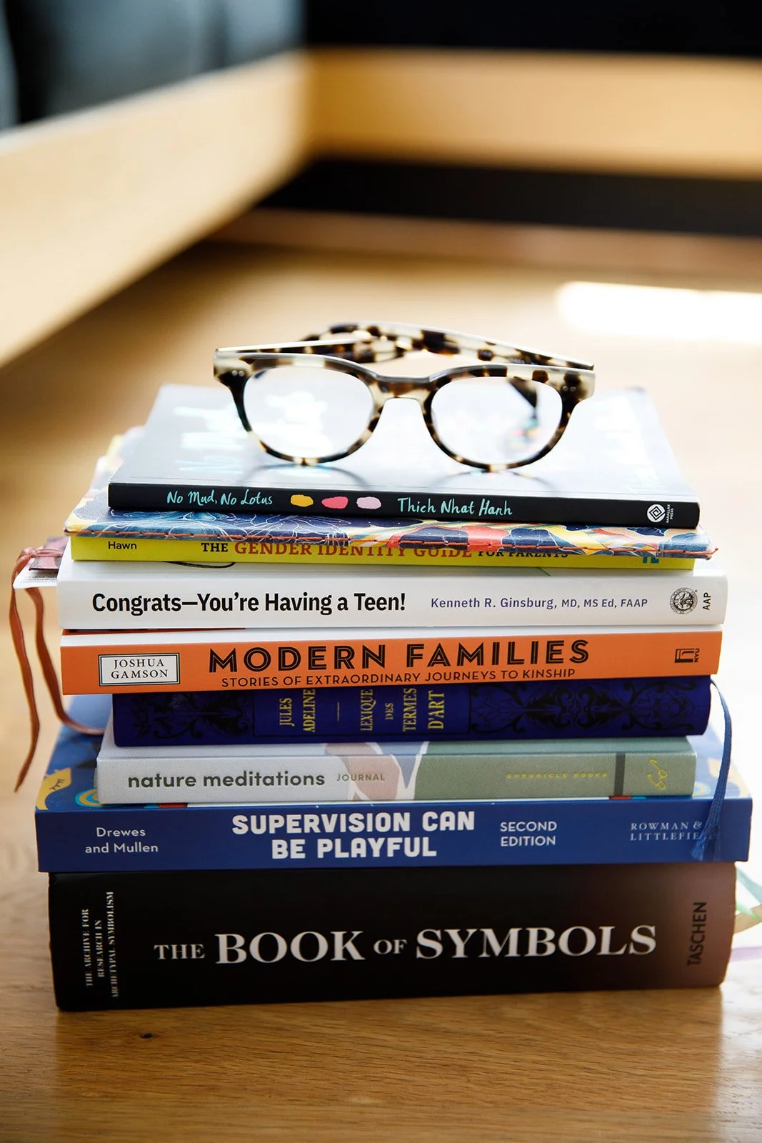 Stack of books on a wooden table with a pair of tortoiseshell glasses on top. The books include topics like gender identity, family stories, meditation, symbols, and supervision.