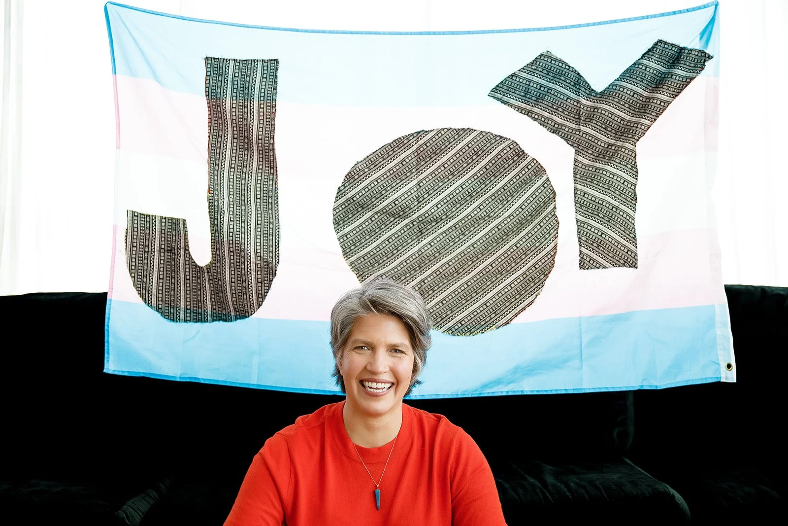 A woman with short gray hair smiling, sitting in front of a flag that has the word "Joy" written on it in large, patterned letters.