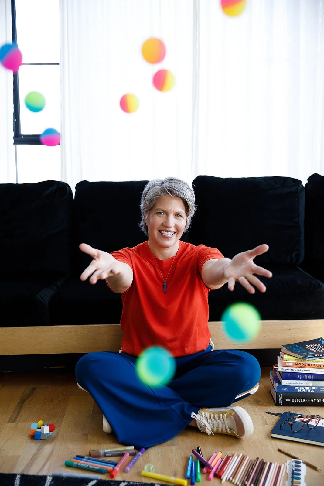 Smiling woman in red shirt and blue pants sitting cross-legged on floor surrounded by art supplies, books, and colorful balls falling from above in a bright room with black couch and white curtains.