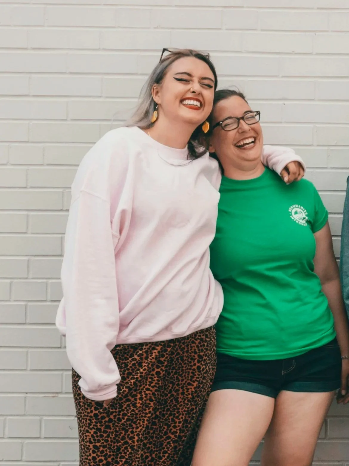 Two women smiling and laughing, standing close together against a white brick wall. One woman is wearing a pink sweatshirt and leopard print pants, and the other woman is wearing a green T-shirt and shorts.