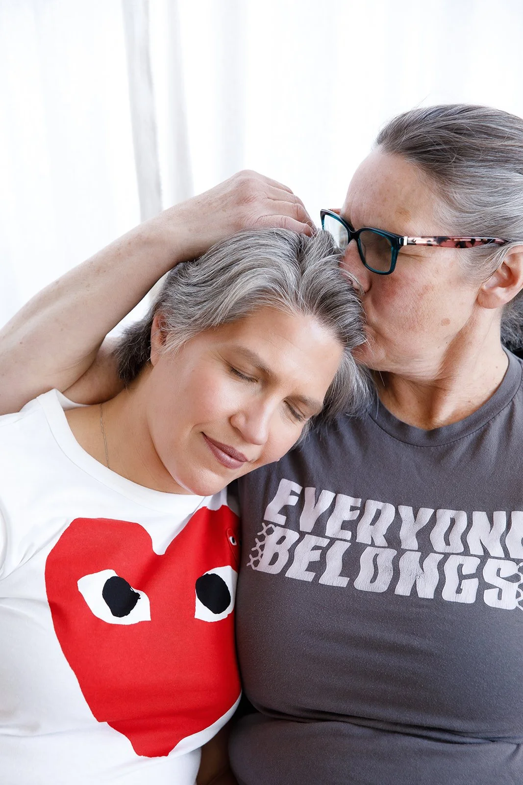 An elderly woman and a middle-aged woman are cuddling, with the elderly woman kissing the middle-aged woman on the forehead as she closes her eyes happily. The elderly woman is wearing glasses and a gray t-shirt, the middle-aged woman is wearing a white t-shirt with a red heart and black eyes design.