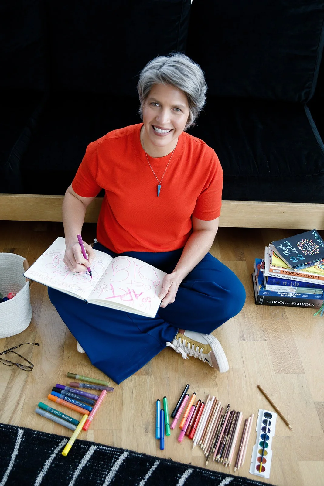 A woman with short gray hair wearing a red shirt and blue pants is sitting cross-legged on a wooden floor, smiling, and drawing with markers in a sketchbook. There are colorful markers, pencils, and art supplies spread out on the floor around her, as well as a stack of books nearby.