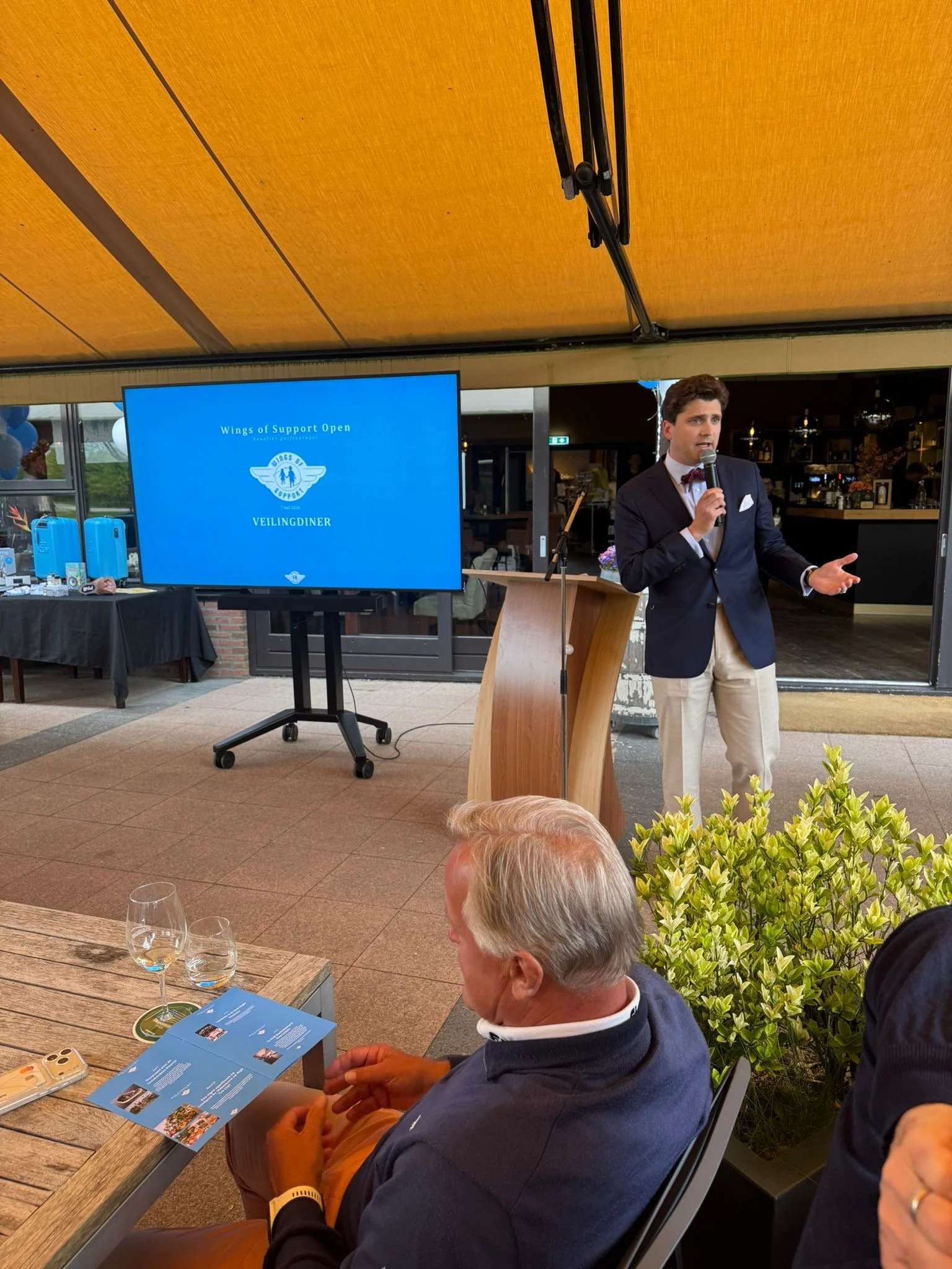 Een man in een pak met een plattegrond op zijn schoot zit aan een tafel tijdens een evenement, met waterglazen en een telefoon voor zich. Op de achtergrond spreekt een andere man met een microfoon, er is een groot scherm met de tekst 'Wings of Suppor