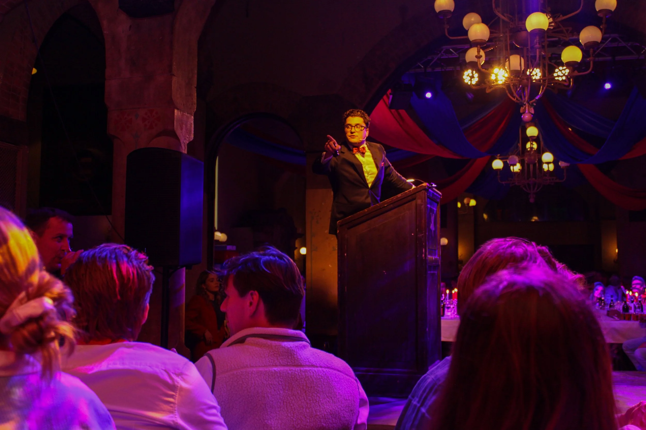 Een man in een zwart pak met een oranje strikboa en een witte overhemd staat op een podium in een donkere zaal met kleurrijk decor en een grote metalen kroonluchter. Hij wijst met zijn vinger naar het publiek, terwijl mensen in de menigte naar hem ki