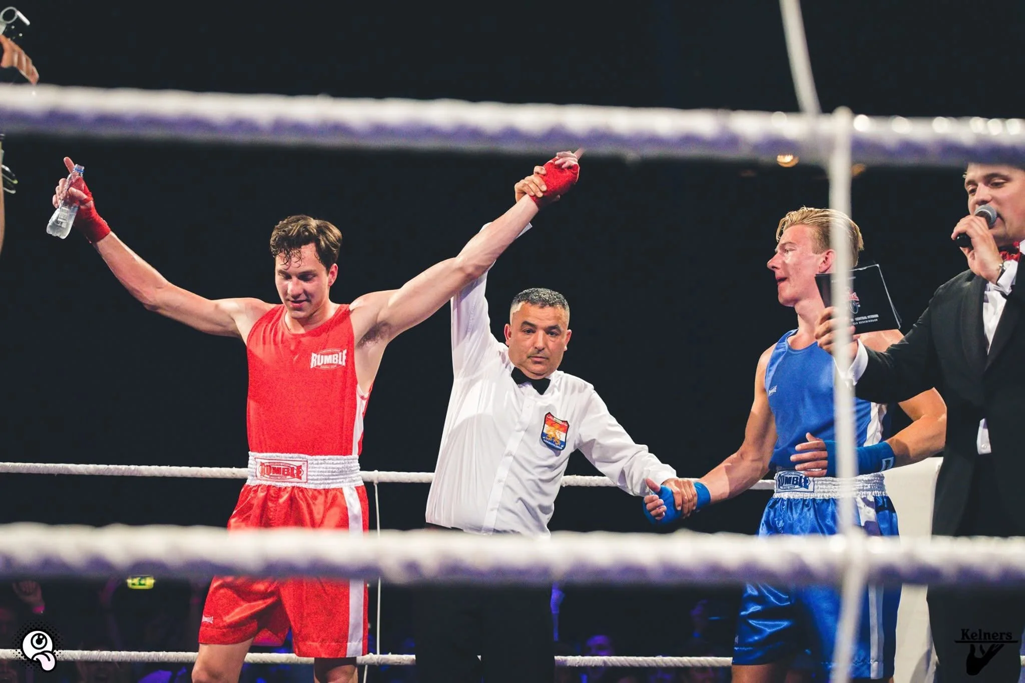 Ringannouncer en een scheidsrechter in de ring, een bokser in rood viert zijn overwinning met opgestoken armen.