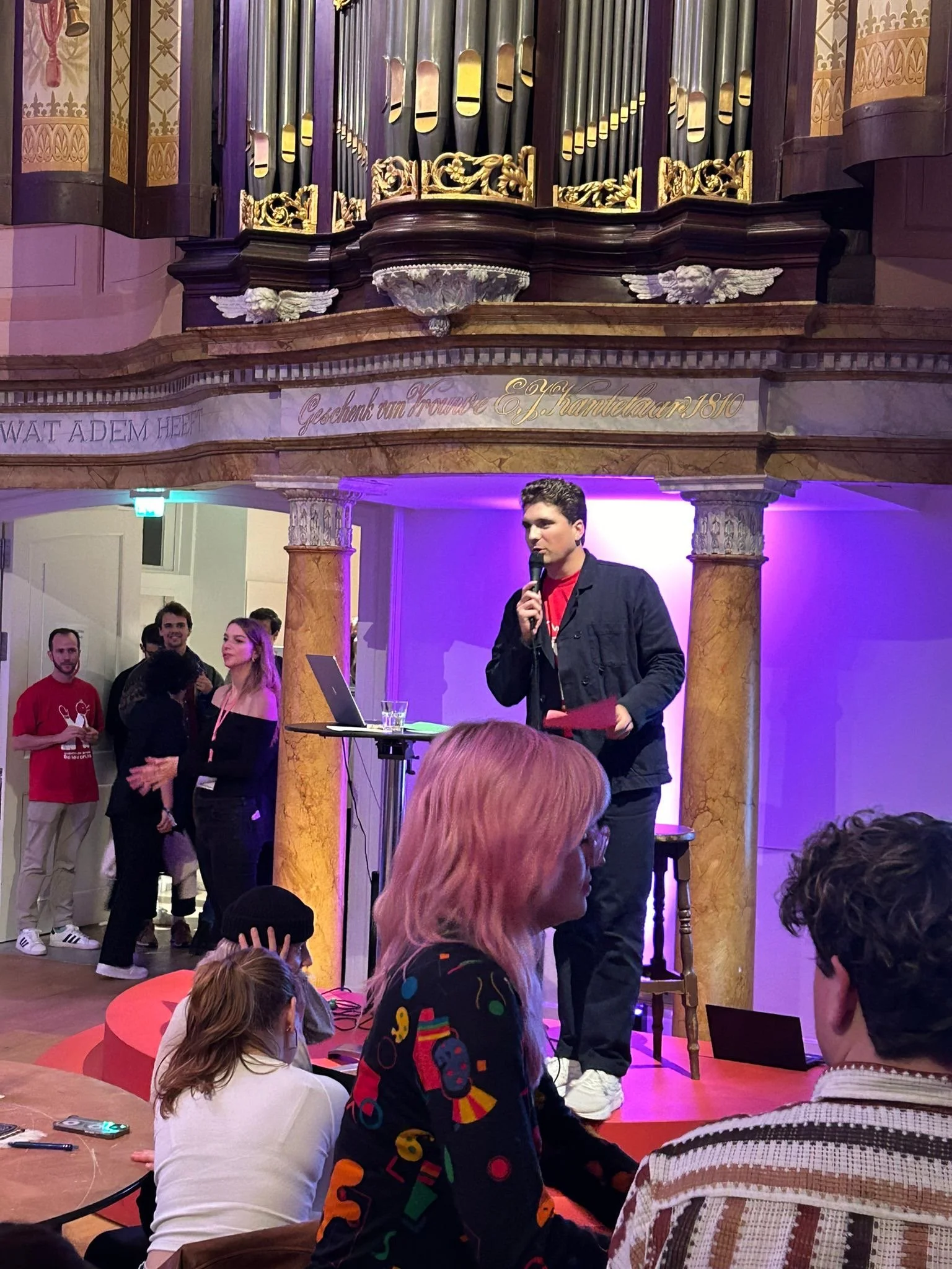 Een man houdt een microfoon vast terwijl hij op een podium spreekt voor een groep mensen in een zaal met klassieke decoraties.