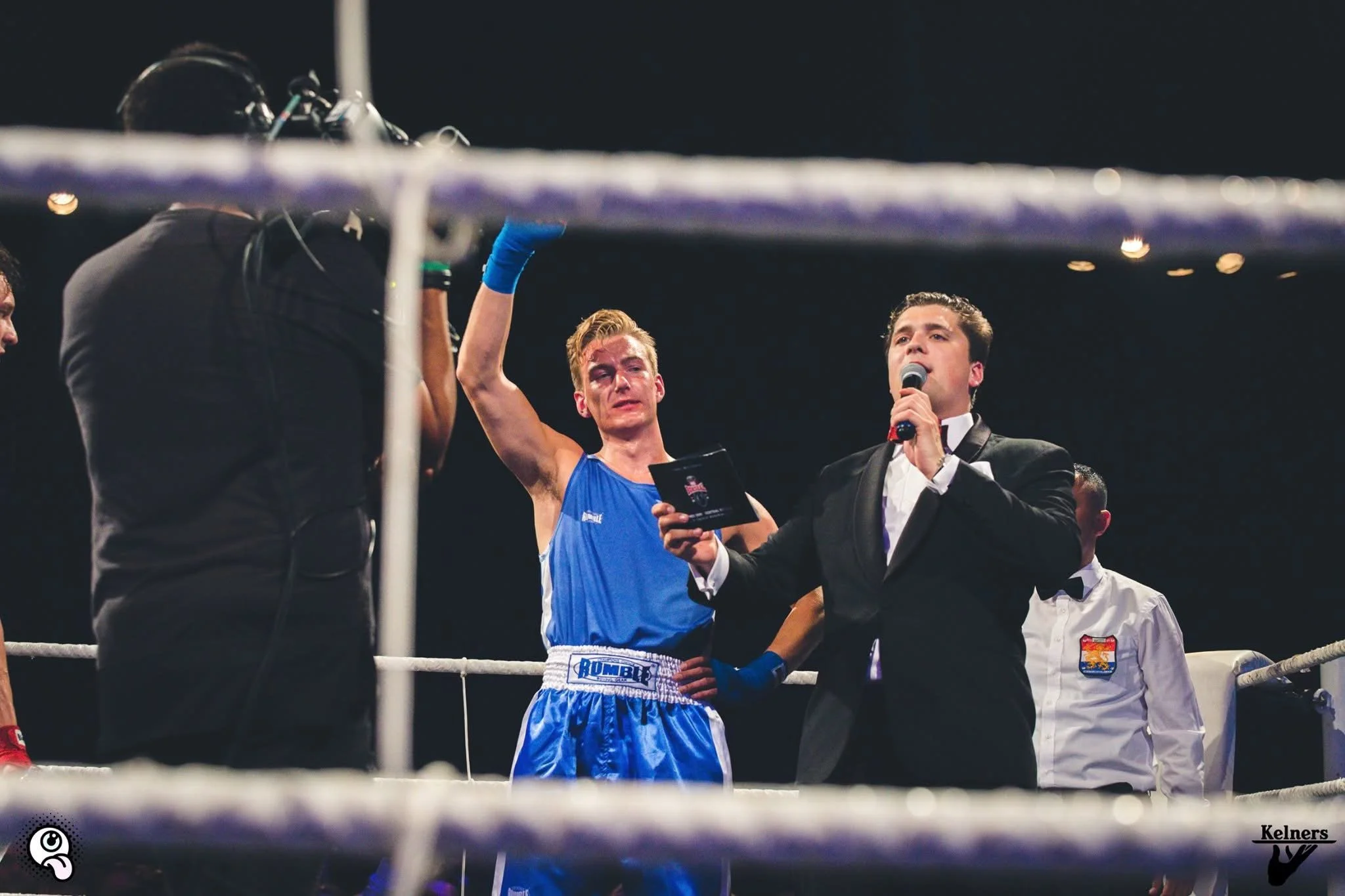 Een man in een blauwe boxoutfit die als bokser in de ring staat, wordt door een presentator in een zwarte pak geïnterviewd terwijl hij een microfoon vasthoudt. Er is een persoon met blauwe bokshandschoenen en een elektronische polshanger. De sfeer is
