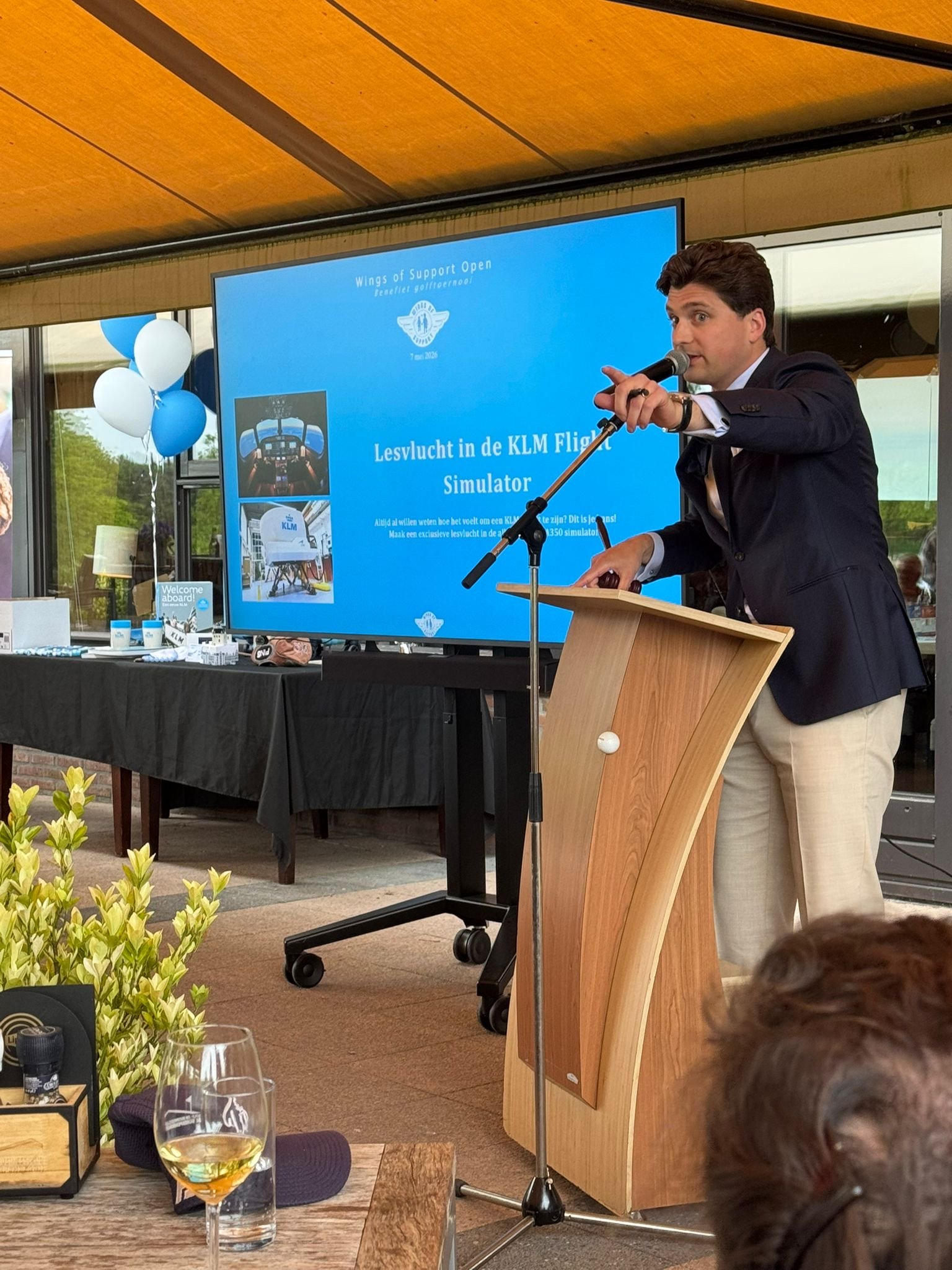 Een man in een donkerblauw jasje spreekt in een microfoon tijdens een presentatie voor een grote monitor. Op de monitor staat de tekst 'Wings of Support Open, Lesvlucht in de KLM Flight Simulator', samen met enkele foto's en het KLM-logo. Op de tafel