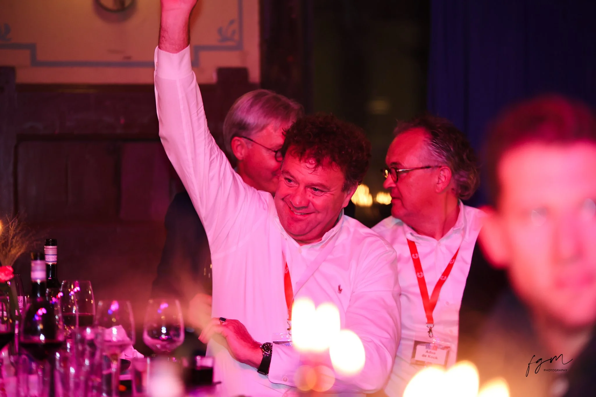 Drie mannen genieten van een feest in een donkere ruimte met pinkkleurige verlichting, wijnflessen en glazen op tafel.
