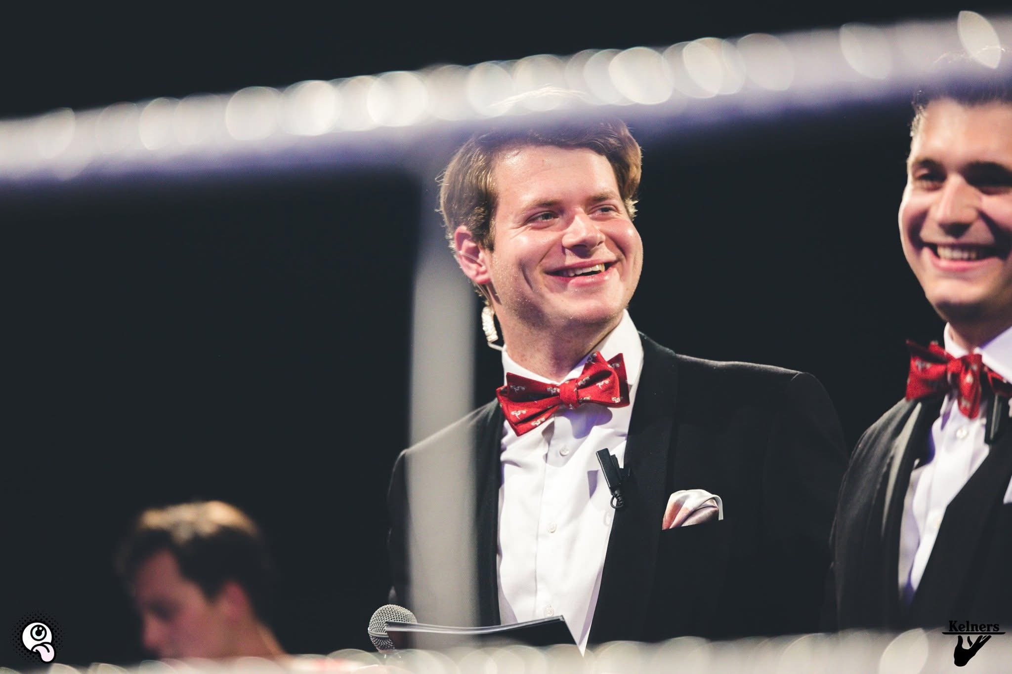 Twee mannen in smoking met rode strik geven een toespraak of presentatie, op een podium met microfoon en achtergrondverlichting.