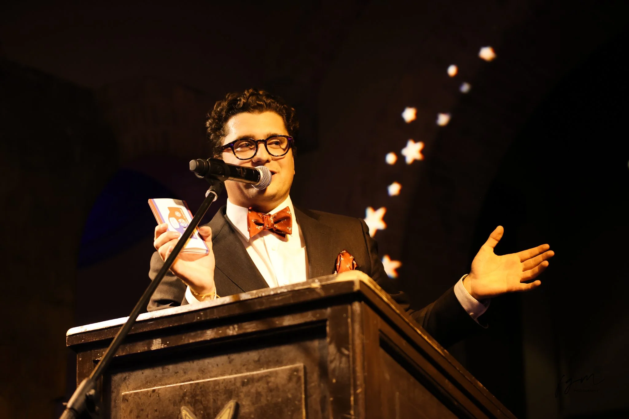 Man in een tuxedo en bril, die spreekt achter een microfoon en een boek vasthoudt, terwijl hij op een podium staat met sterrenverlichte achtergrond.