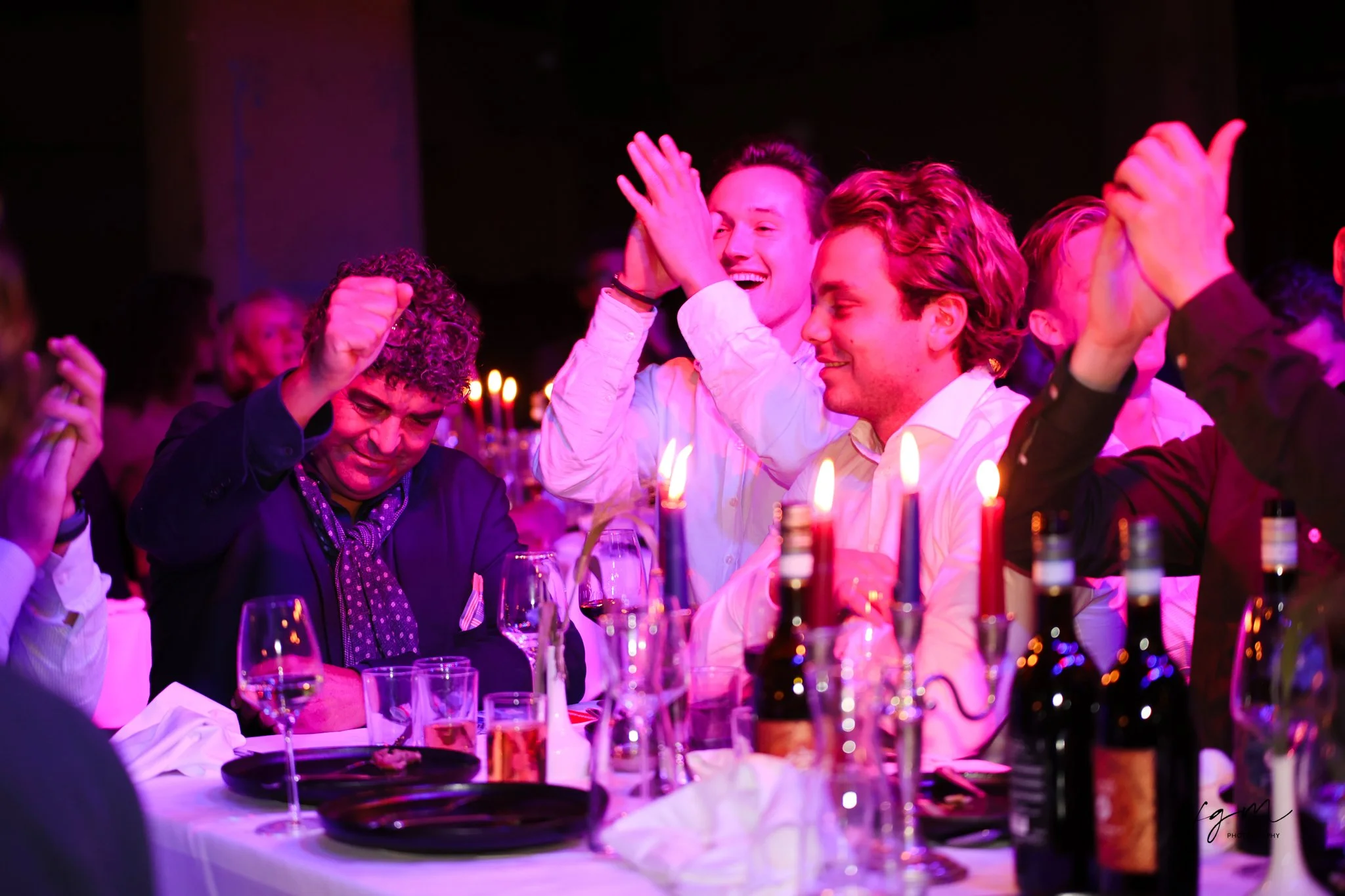 Mensen die lachen en plezier maken tijdens een feestelijke gebeurtenis, omringd door wijn en kaarslicht in een donkerverlichte ruimte.