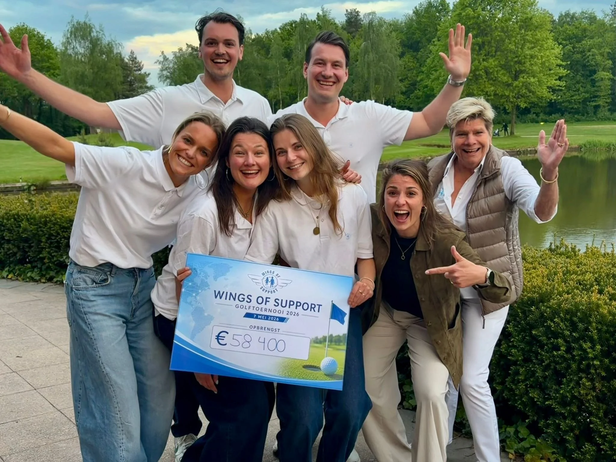 Vijf mensen glimlachend en juichend, twee vrouwen houden een groot cheque van 58.400 euro voor Wings of Support. Ze staan buiten in een park met groen erfgoed en een meer op de achtergrond.