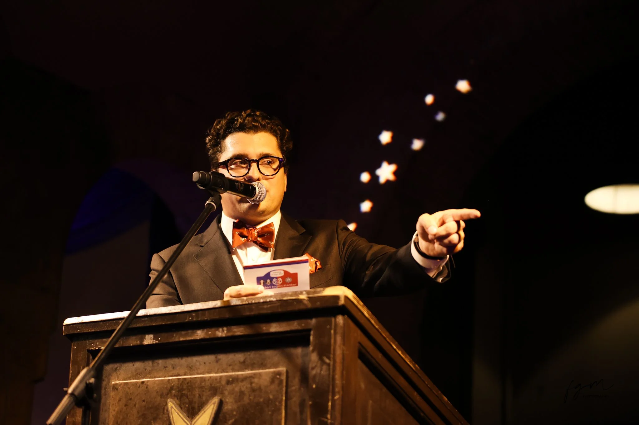 Man in smoking en bril, spreekt achter een ouderwetse microfoon, met een korte tekst in zijn hand, op een podium met een boekenkast op de achtergrond.