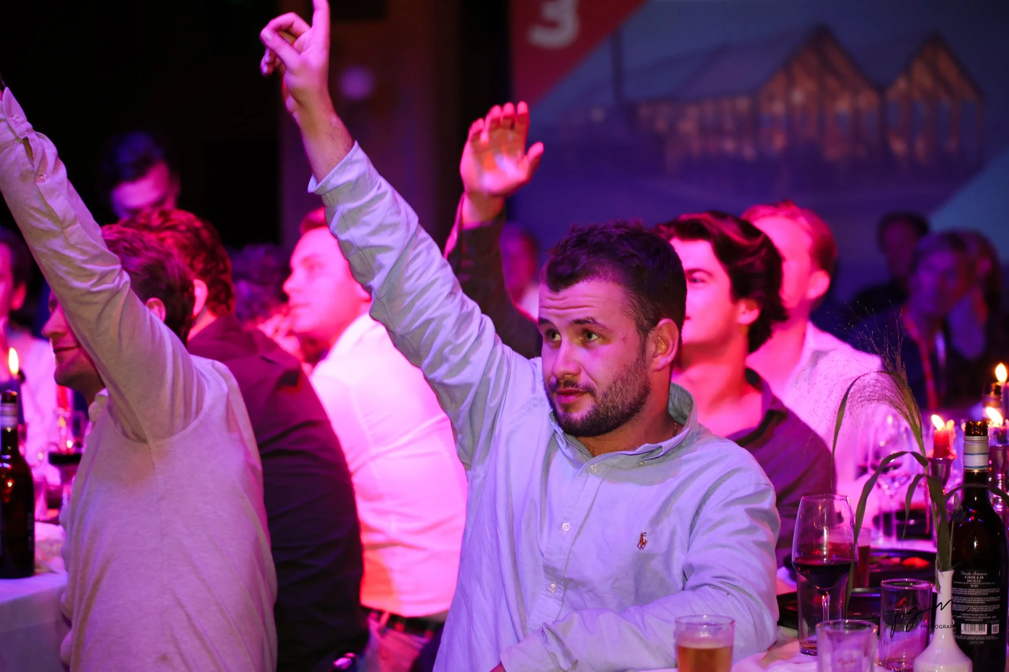 Mannelijke persoon met een lichtblauw overhemd die omhoog wijst tijdens een bijeenkomstd door pinke en blauwe verlichting, omringd door andere mensen aan een tafel met drankjes.