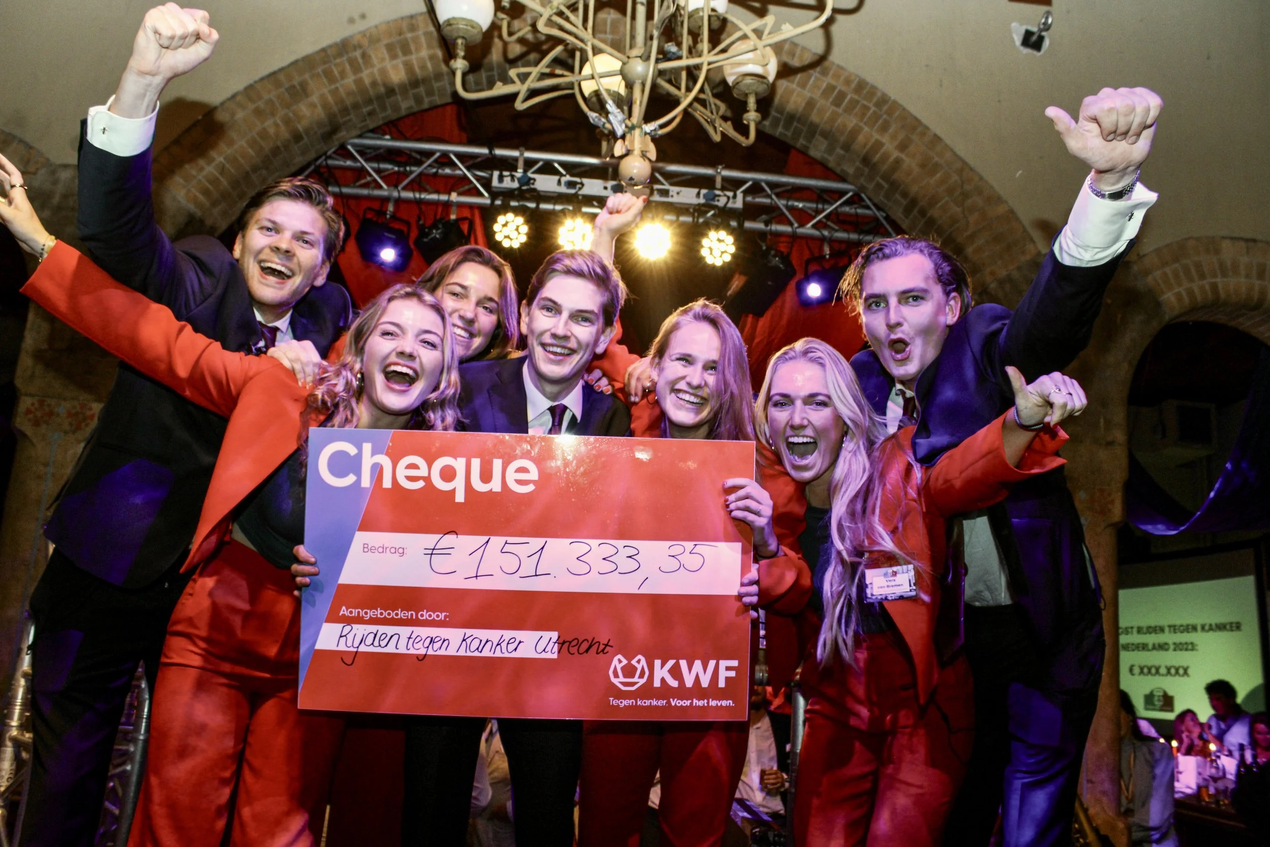 Groep mensen die feest vieren met een groot cheque ter waarde van €151.333,35 voor kankeronderzoek, onder een feestelijk versierde omgeving met lampen en kleurrijke verlichting.