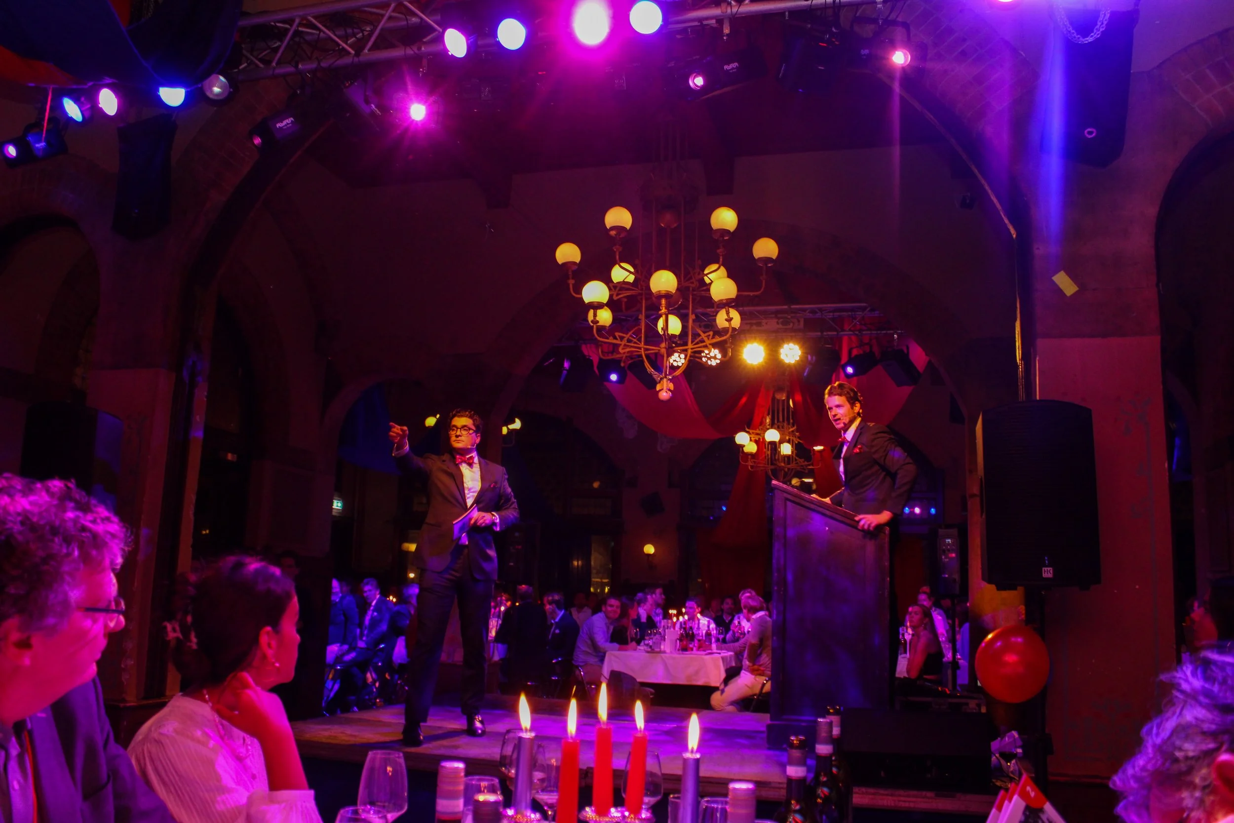 Gezellig feest in een zaal met kleurrijk licht en een kroonluchter, een man en een vrouw staan op het podium terwijl het publiek kijkt.