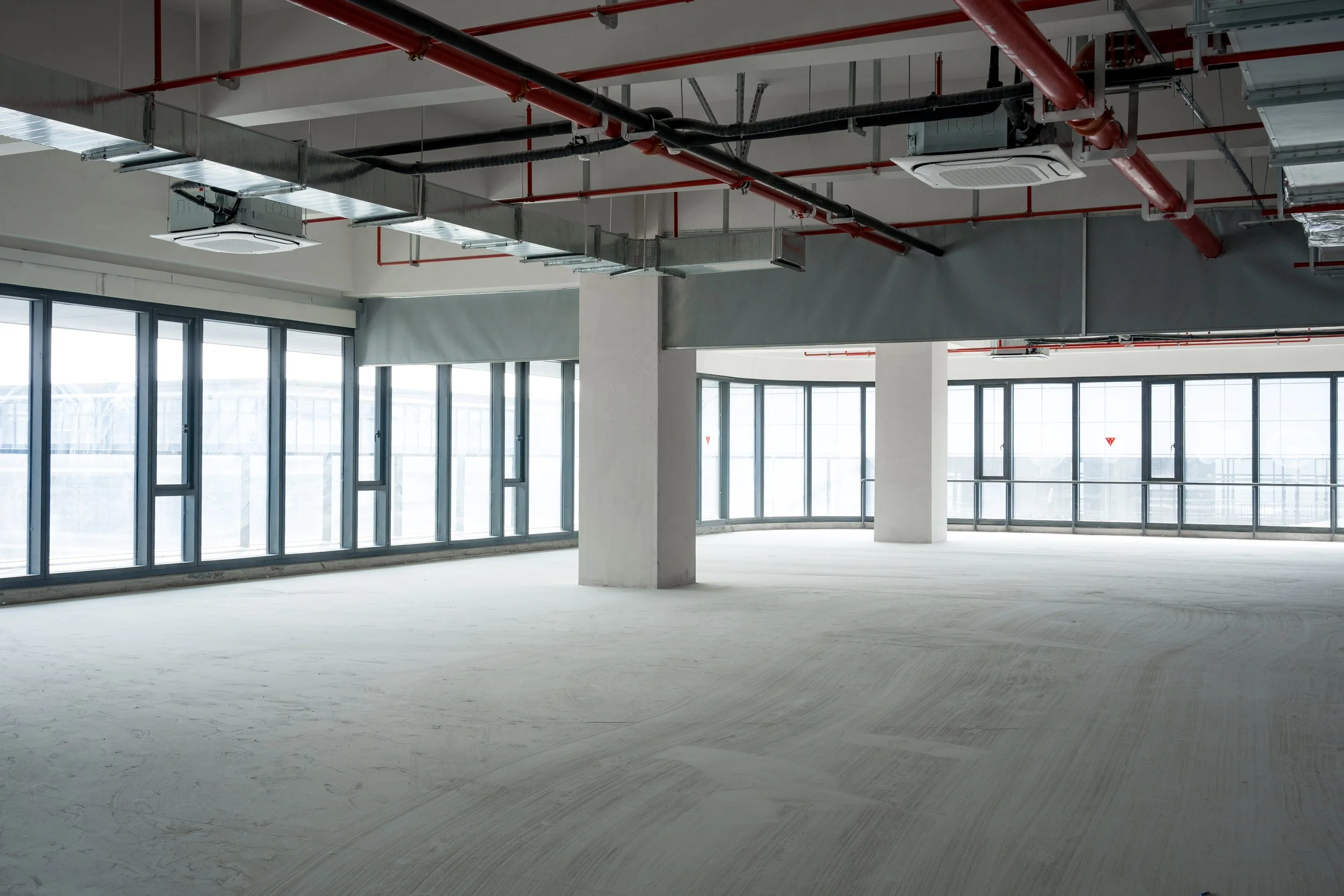 A large, empty commercial space under construction with freshly painted gray and white walls, exposed ceiling ductwork and red piping, concrete support columns, and floor-to-ceiling windows along the perimeter.