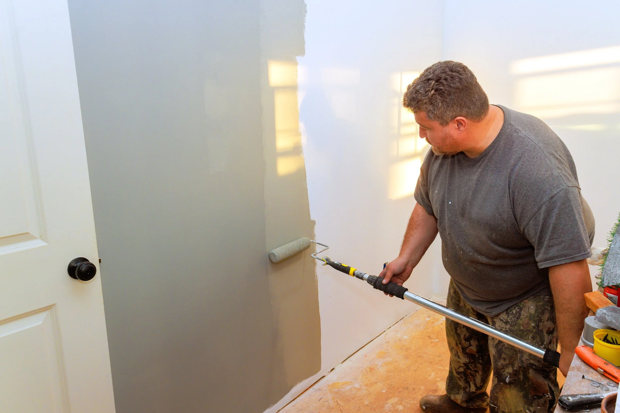 A painter in a paint-stained t-shirt and cargo pants rolling gray paint onto an interior wall next to a white door.