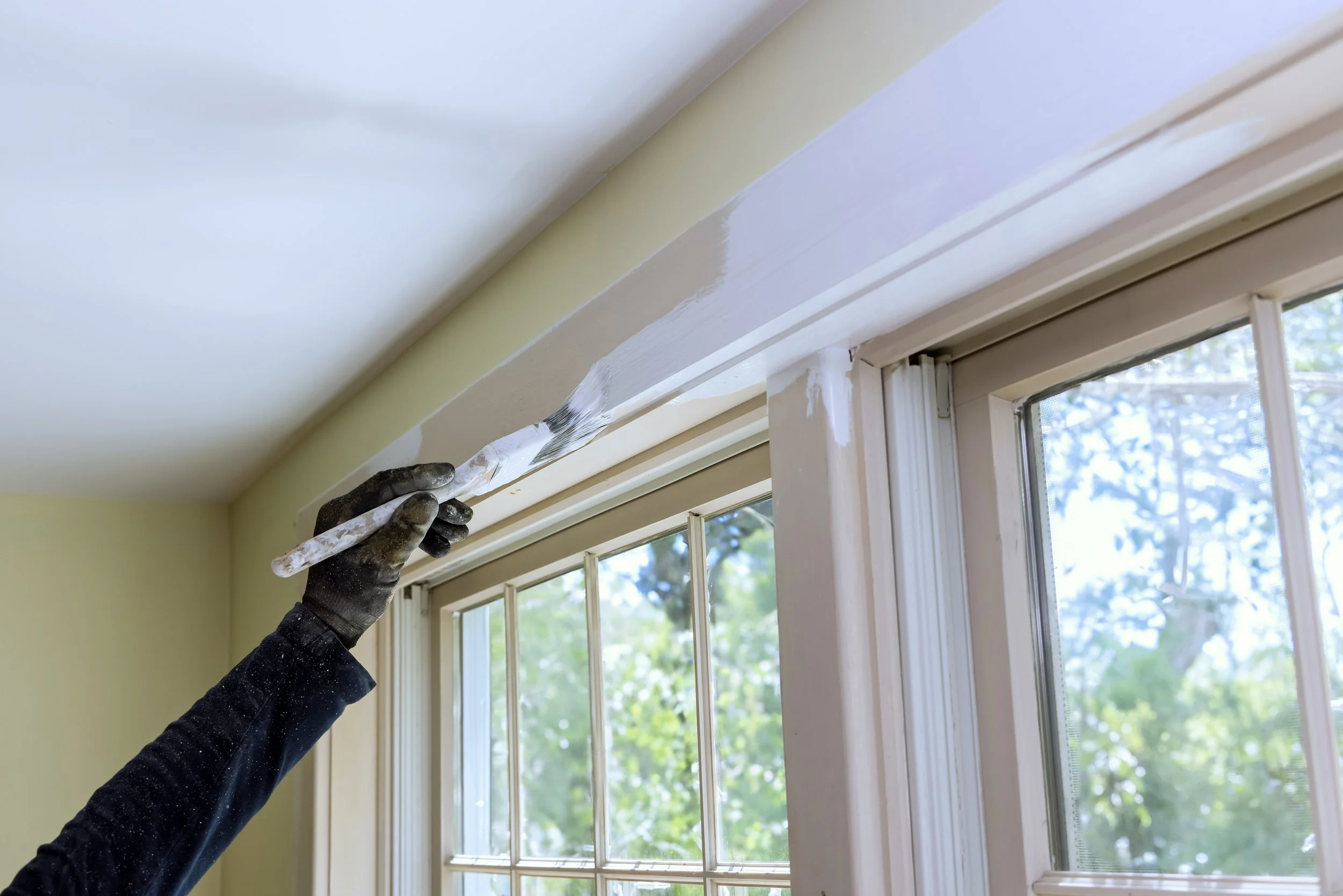 A gloved hand carefully cutting in white paint along the trim above a large interior window using a paintbrush, with light green walls and a view of trees outside.