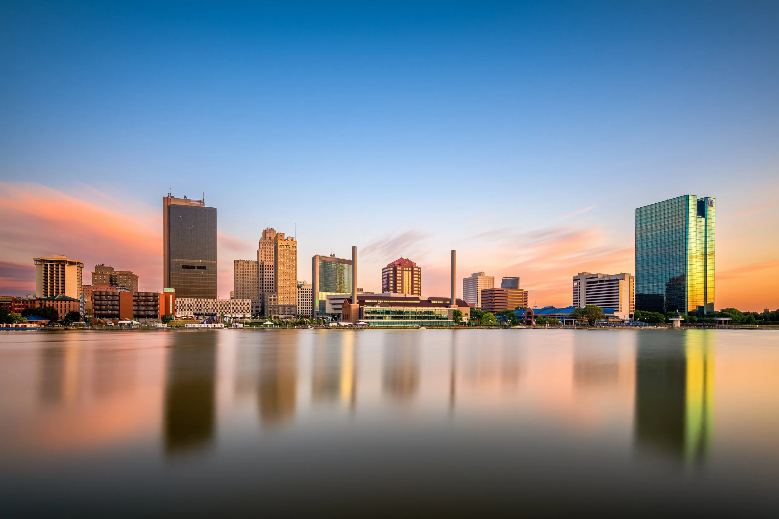 Toledo Ohio city skyline at sunset reflected on the Maumee River, showing downtown buildings and colorful sky