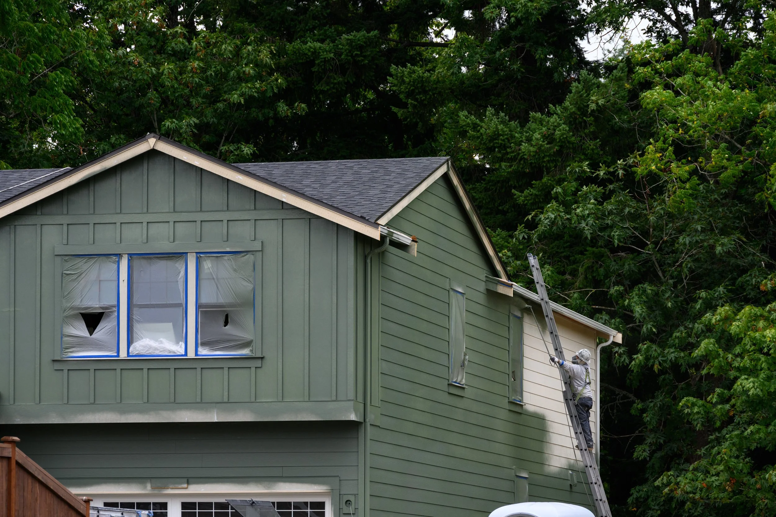 Professional painter wearing a hard hat and respirator on a ladder using a sprayer to paint the exterior of a green house.
