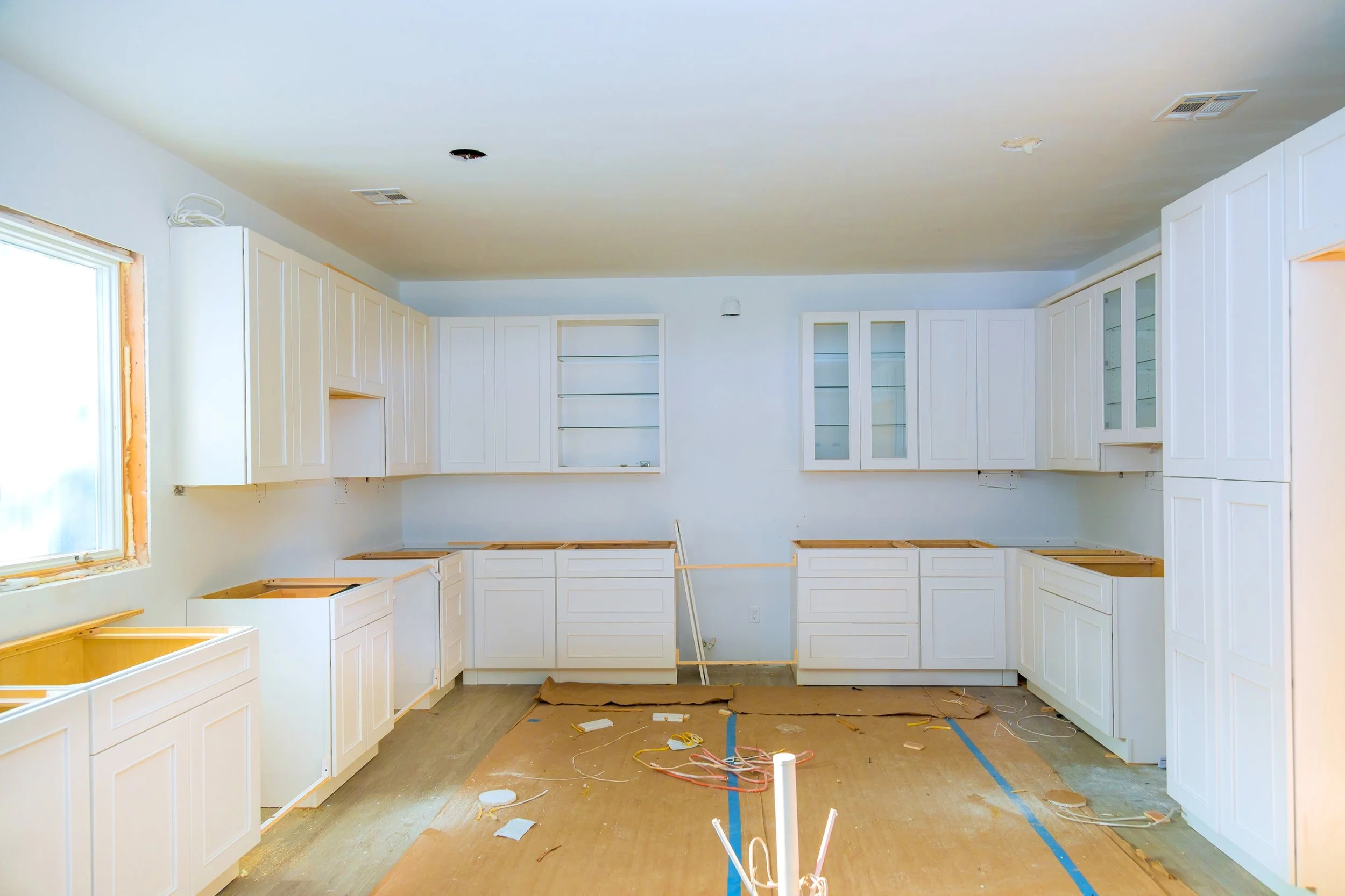 A kitchen under renovation with newly installed white shaker-style cabinets awaiting finishing touches, bare wood countertops, and an unfinished floor covered with construction debris — staged and ready for interior painting.