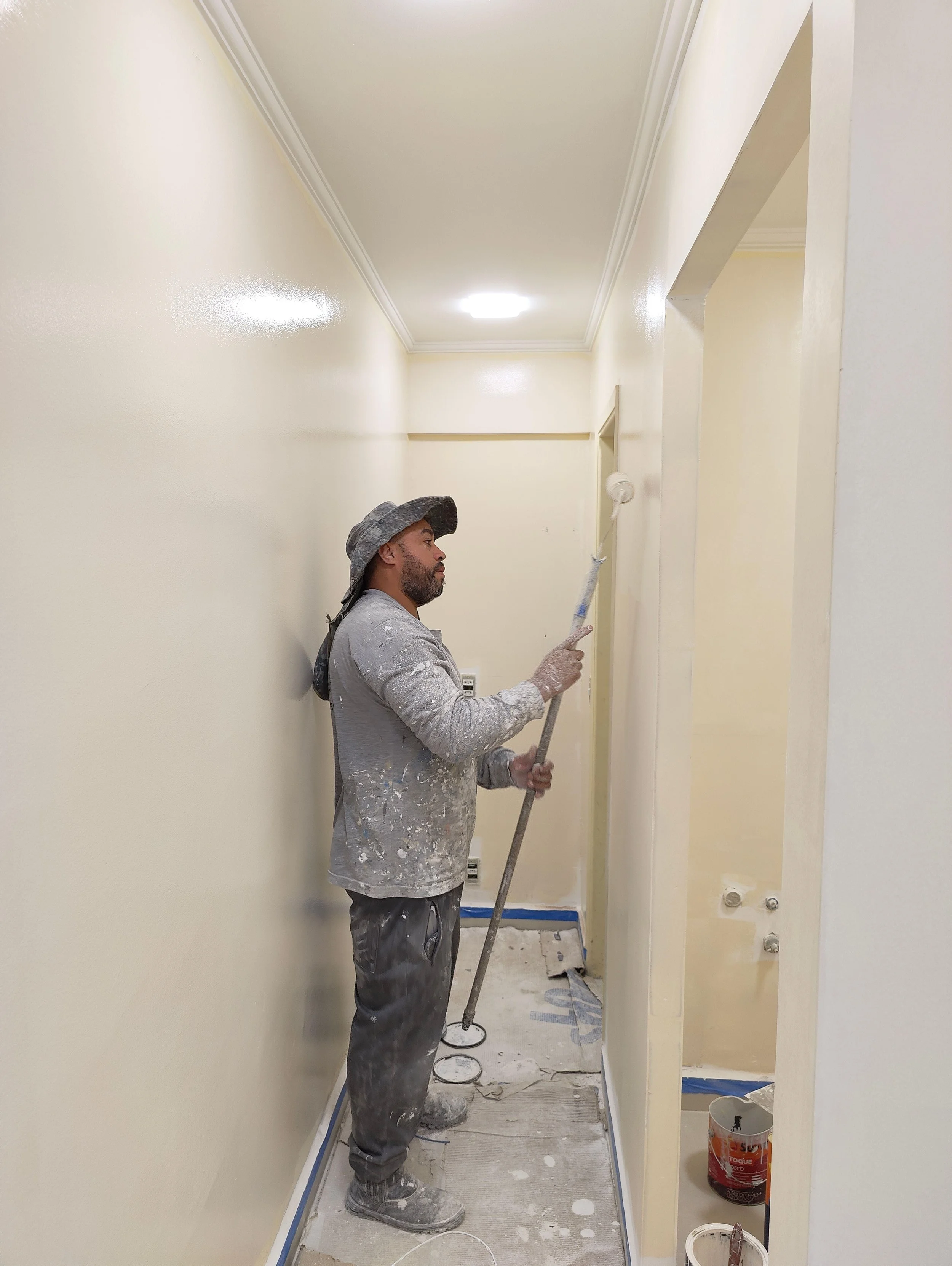 A painter in paint-spattered gray work clothes and a bucket hat rolls white paint onto the wall of a narrow hallway, with paint cans and blue painter's tape visible on the floor.