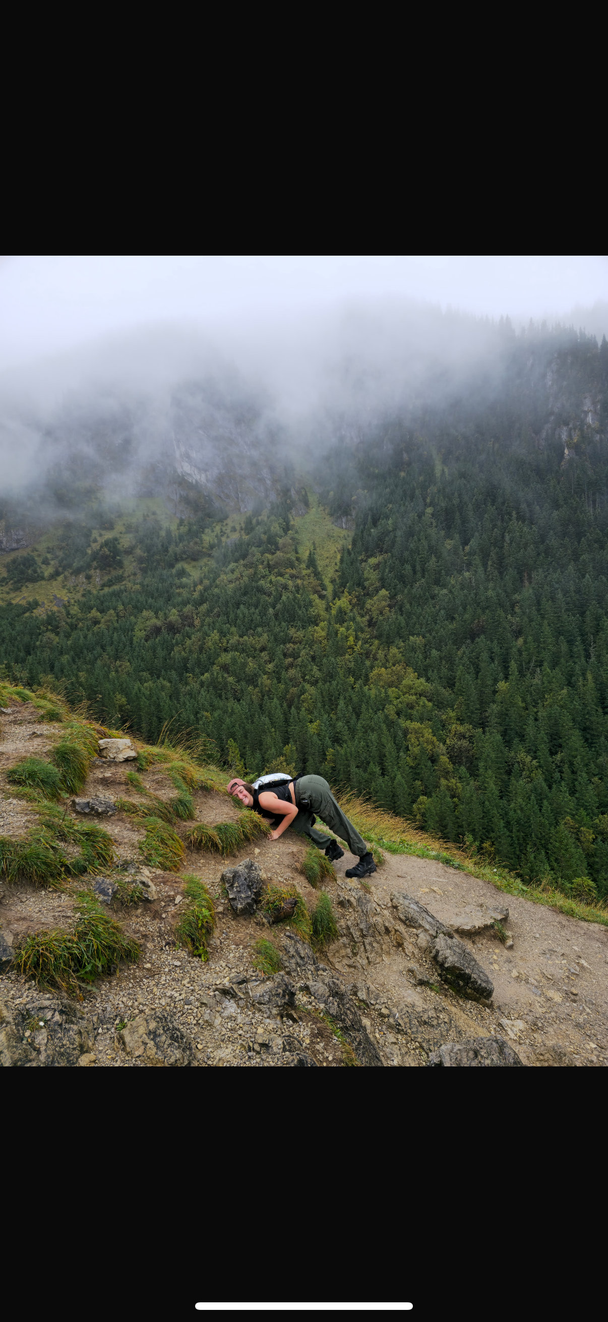 A person crouching on a mountain trail, with lush green forests and misty mountains in the background.