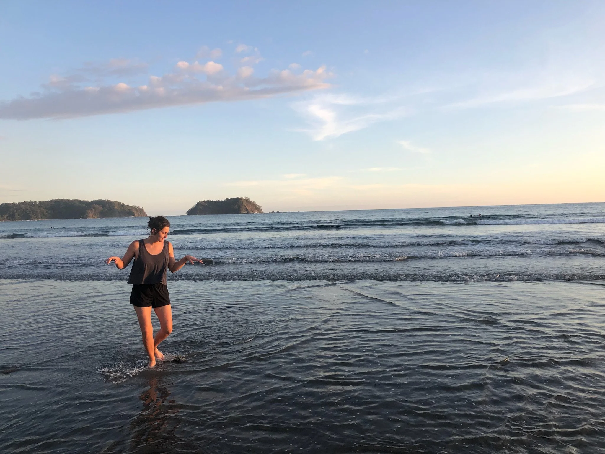 A woman in a black tank top and shorts standing in wet sand at the beach during sunset, with water reaching her knees. In the background, there are small islands or land formations in the ocean, and a few people swimming or playing in the water. The sky is partly cloudy with soft pastel colors from the setting sun.