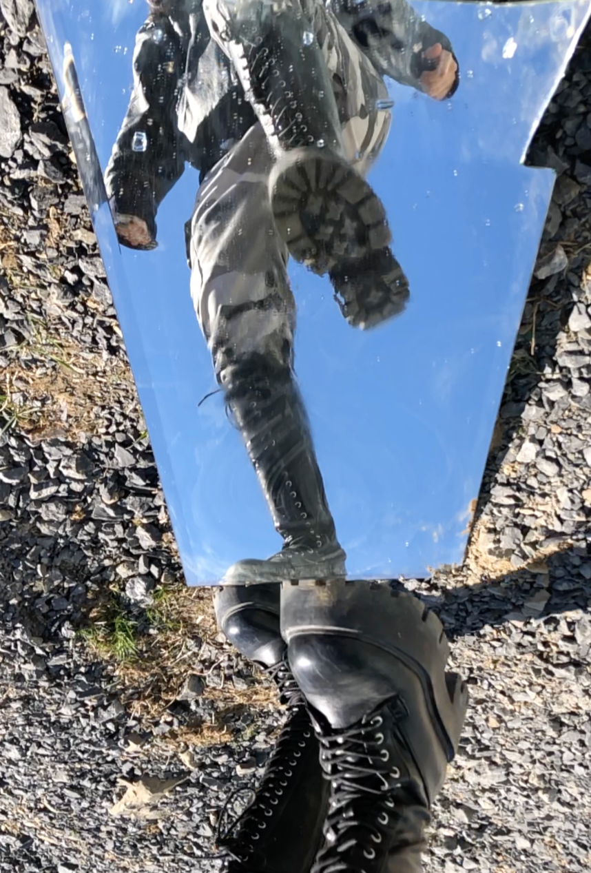 Reflection of person wearing black boots and black clothing in a mirror placed on a gravel surface, with a blue sky reflected in the mirror.