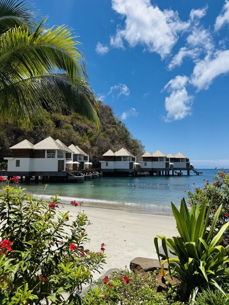 Overwater bungalows with straw roofs on a tropical beach with white sand, lush green plants, and a clear blue sky with a few clouds.