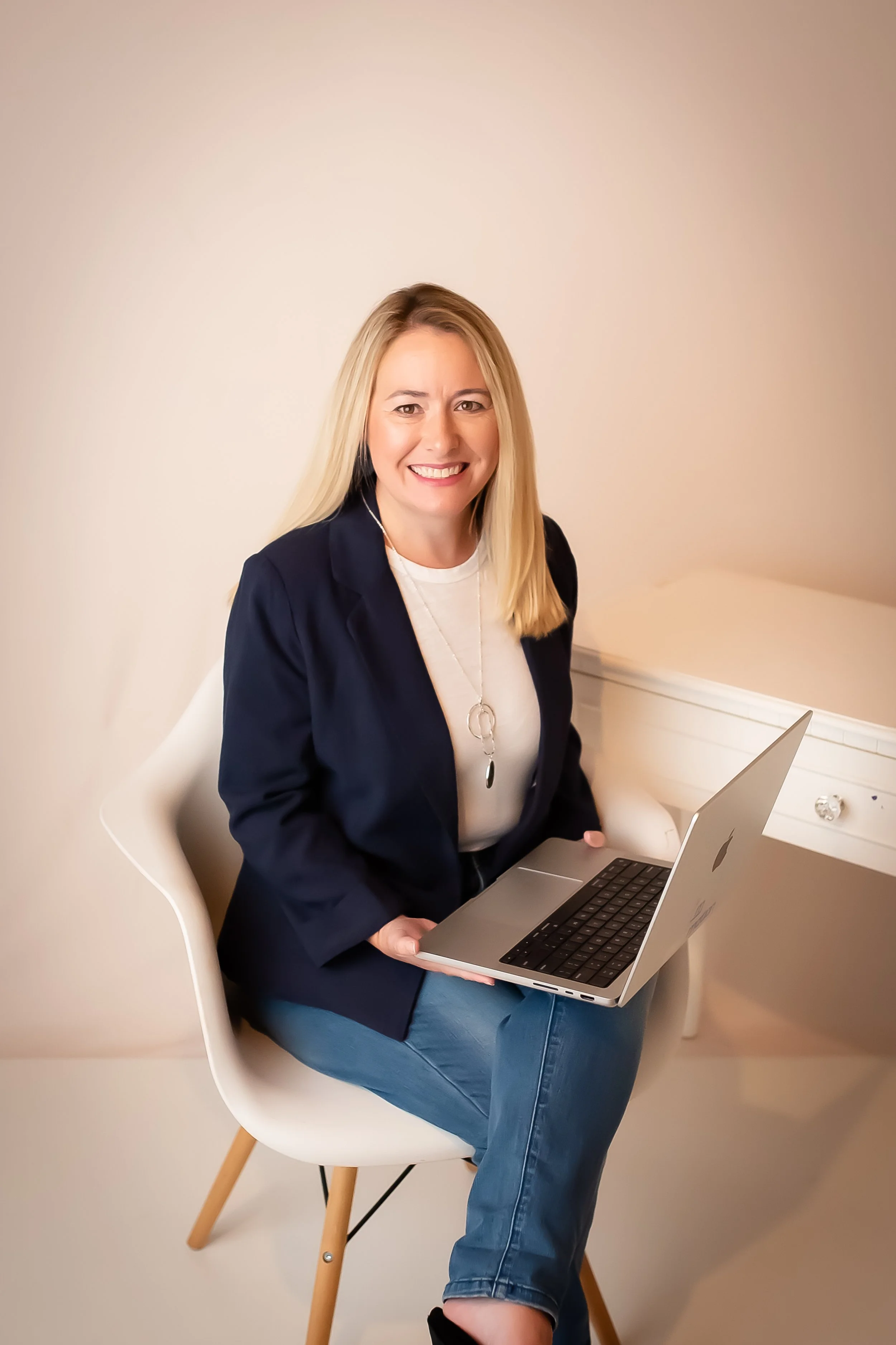 A woman sitting in a white chair, smiling, holding a silver laptop, wearing a navy blazer, white top, and blue jeans, with a neutral beige background.