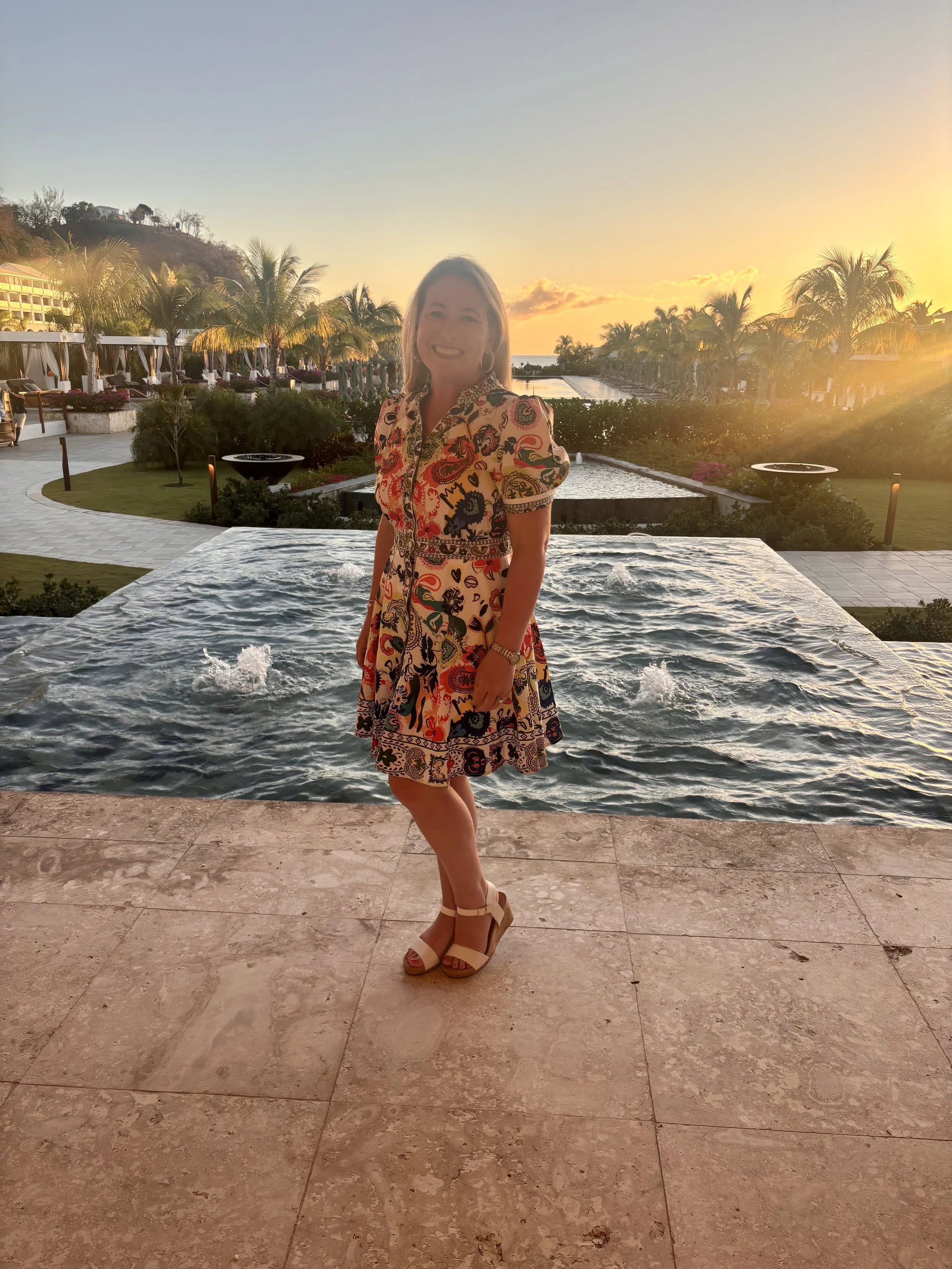 A woman stands on a marble patio in front of a pool with a sunset in the background. She is wearing a colorful, patterned dress and beige high heels, smiling at the camera.