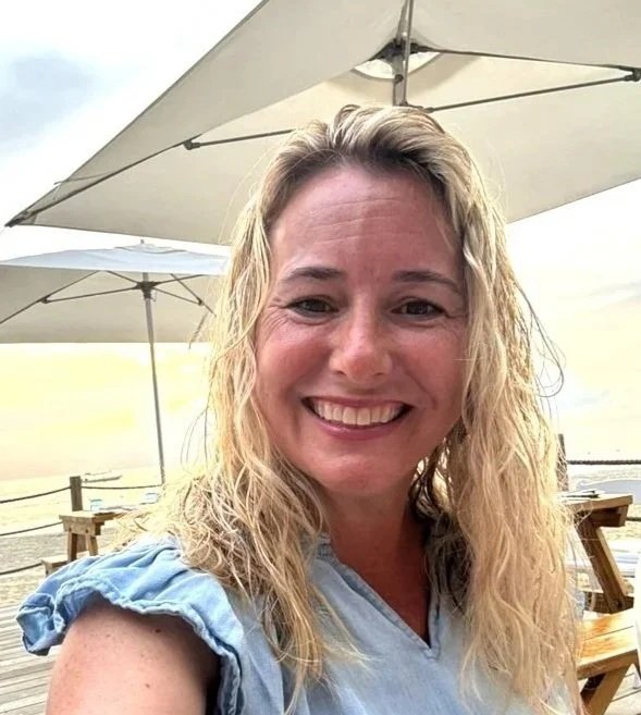 A woman with blonde, wavy hair smiling at the camera outdoors, with large beach umbrellas and tables in the background, on a cloudy day.