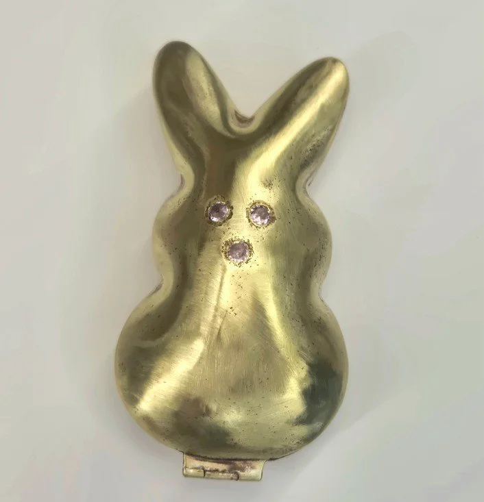 Gold-colored bunny-shaped object with three small pink gemstones on its face against a light background.