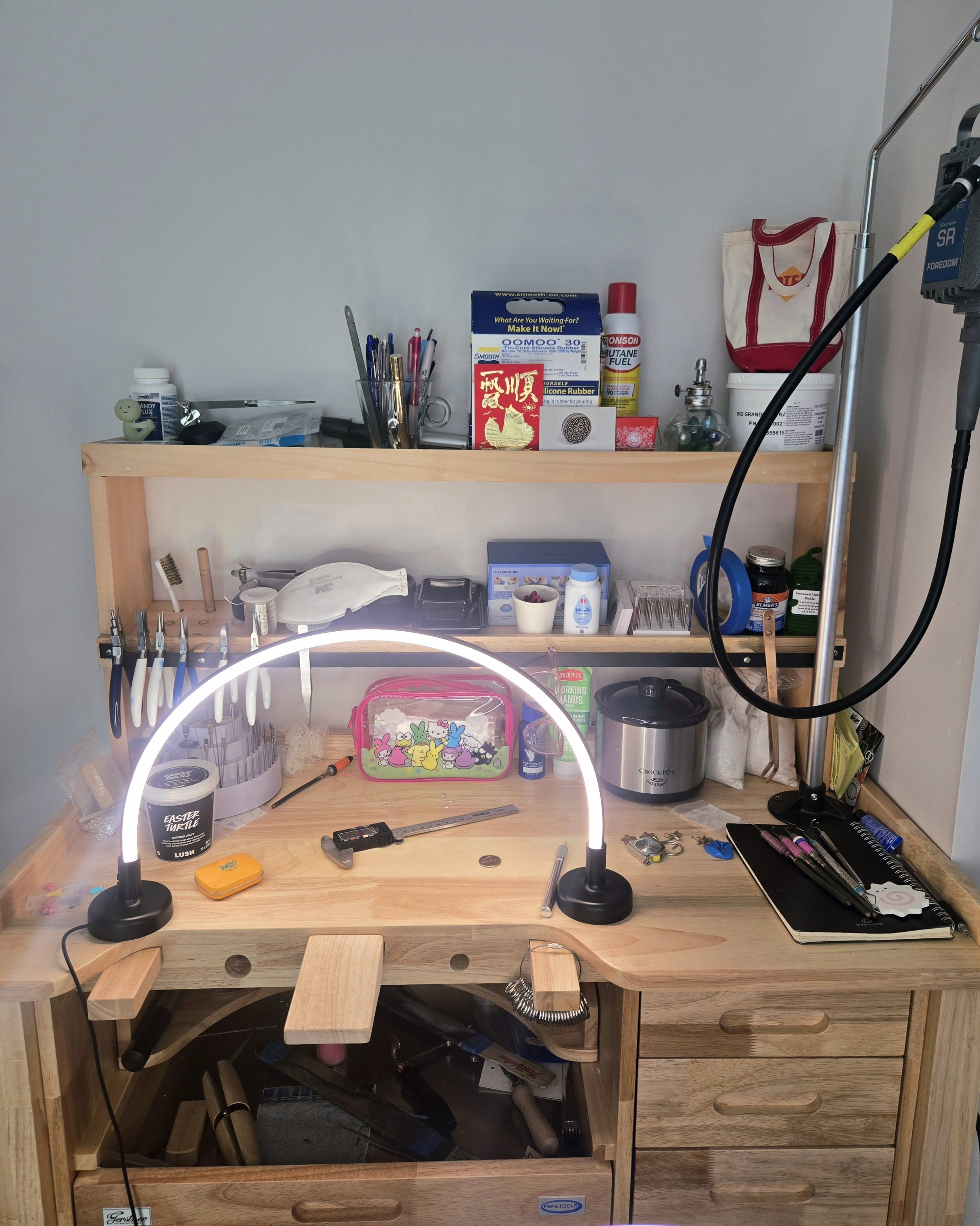 A wooden lighted jewelry with multiple compartments holding various small items, tools, and containers. On the left side, there are drill-like tools, and in the middle, there is a pink cartoon-themed pencil case, a ring, and other small objects. To the right, there are bottles, a black notebook, and a few pens on a tray. Below the organizer, a lower shelf stores additional tools and supplies, including a box and some boxes of items.