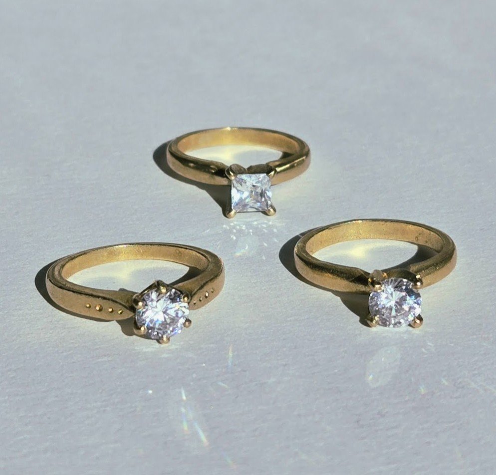 Three gold rings with diamond stones on a white surface, one with a princess cut diamond, and two with round brilliant cut diamonds.