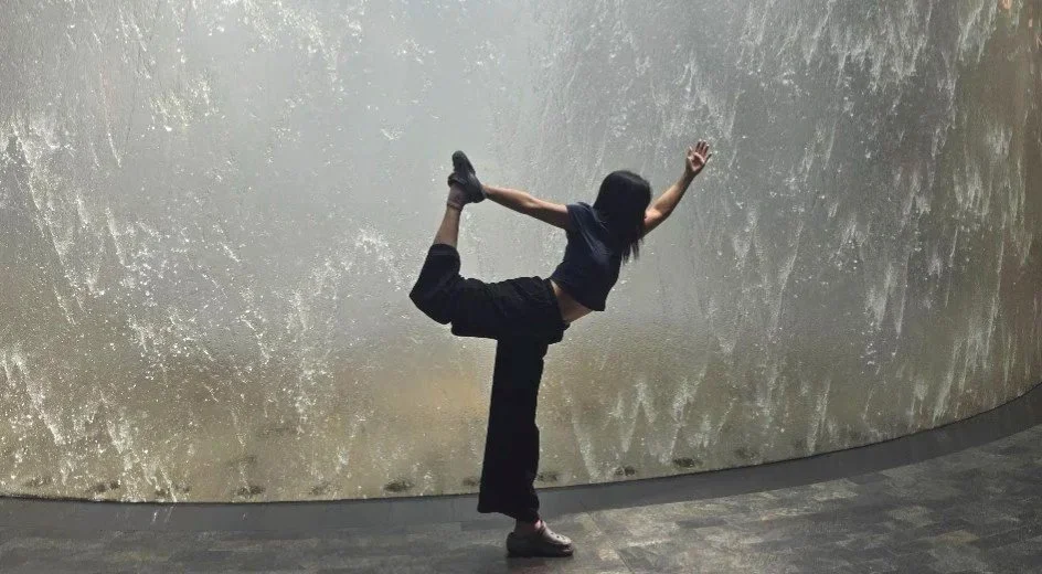 A girl in a black outfit doing a balancing dancer pose with one leg bent behind her and arms extended, standing in front of a large waterfall at Singapore airport. Vortex waterfall Changi airport.