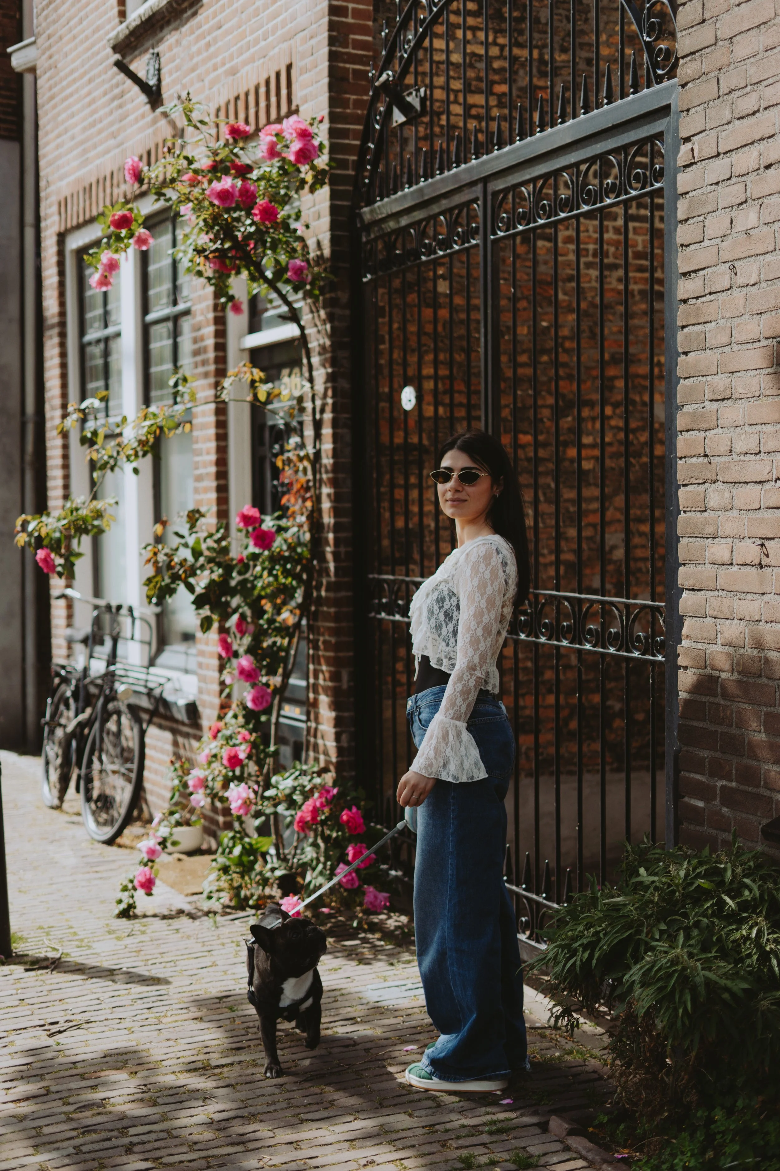 A woman with dark hair wearing glasses, a white lace long-sleeve top, and blue jeans standing on a cobblestone street holding a leash attached to a black and white dog. There are pink flowers and bicycles on the sidewalk, and a brick building with a 
