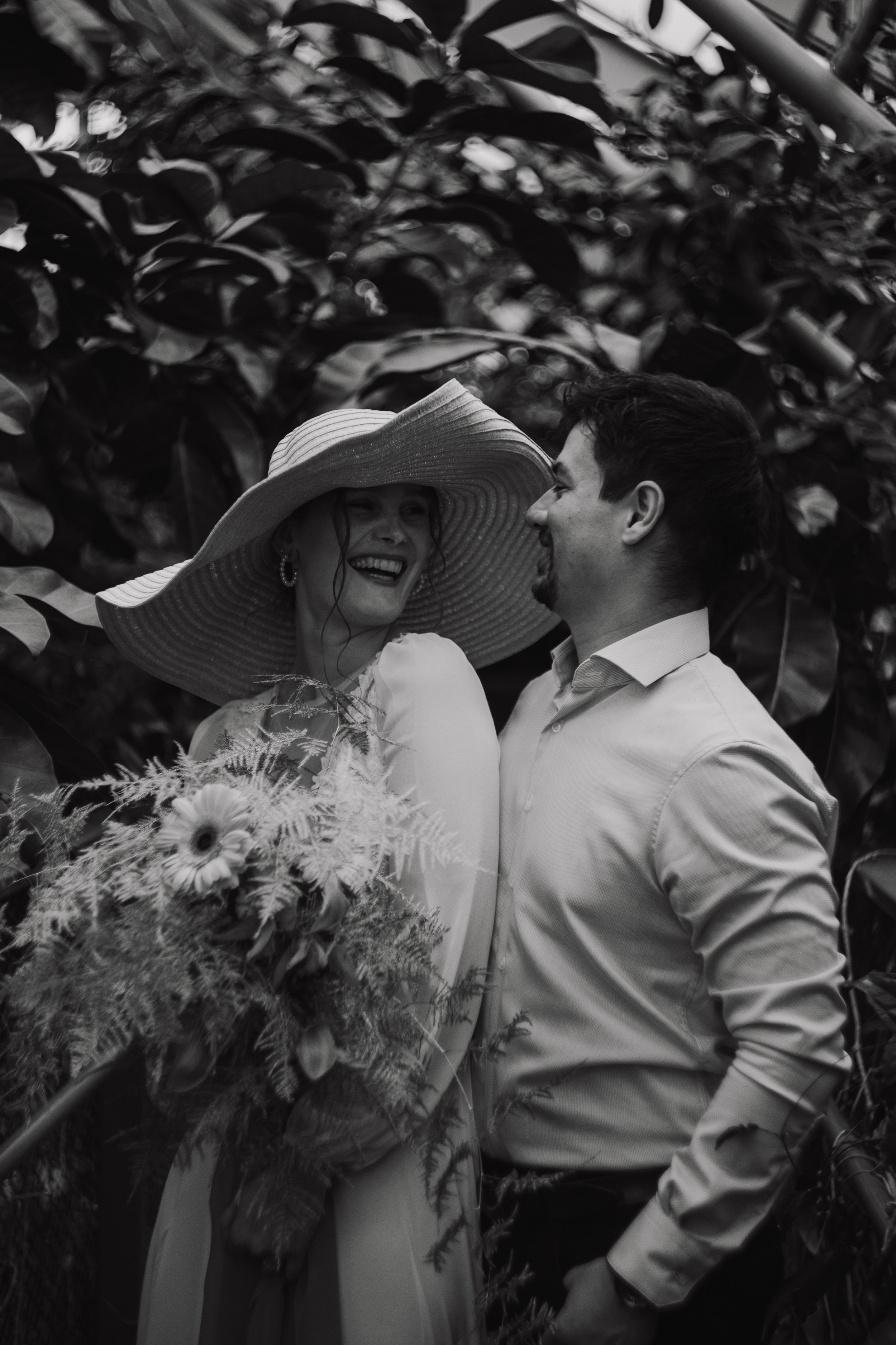 A couple with light skin smiling at each other in front of dense foliage. The woman wears a wide-brimmed hat and dress, holding a bouquet of flowers. The man wears a light-colored shirt, facing her.