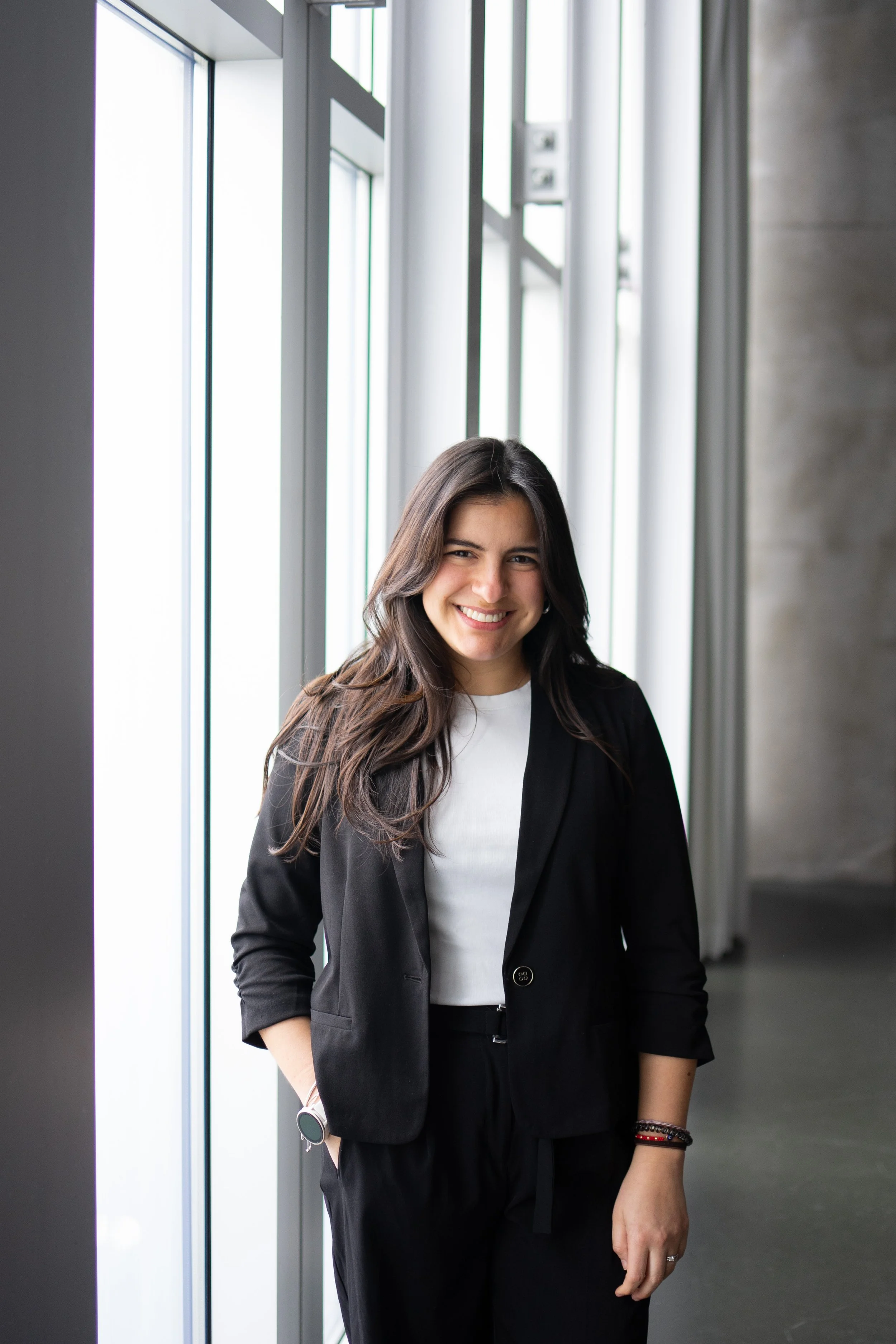 A young woman with long brown hair smiling, dressed in a black blazer and white shirt, standing indoors near large windows.