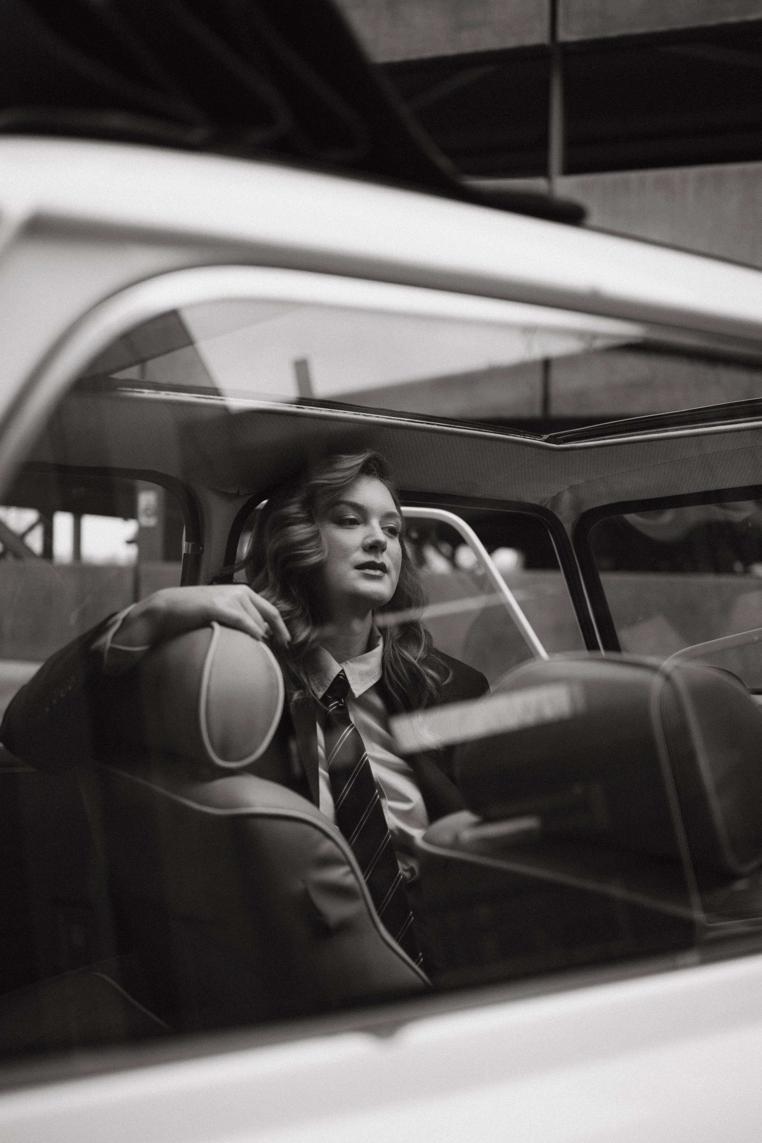 A woman with wavy hair sitting in a vintage car, resting her arm on the window frame, with a calm expression. The image is in black and white.