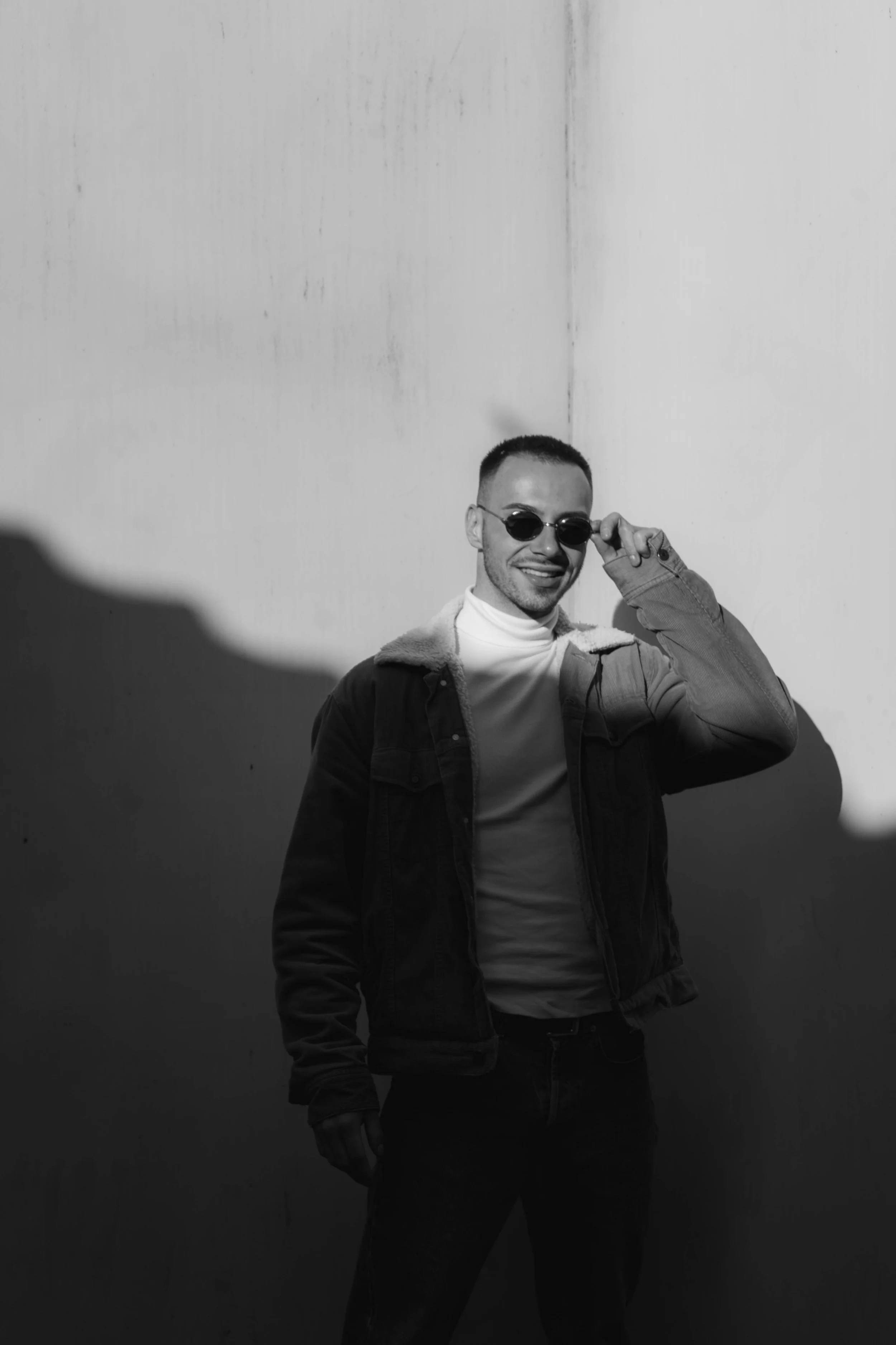 A young man wearing sunglasses, a light-colored turtleneck, and a denim jacket smiling at the camera, standing against a plain wall.